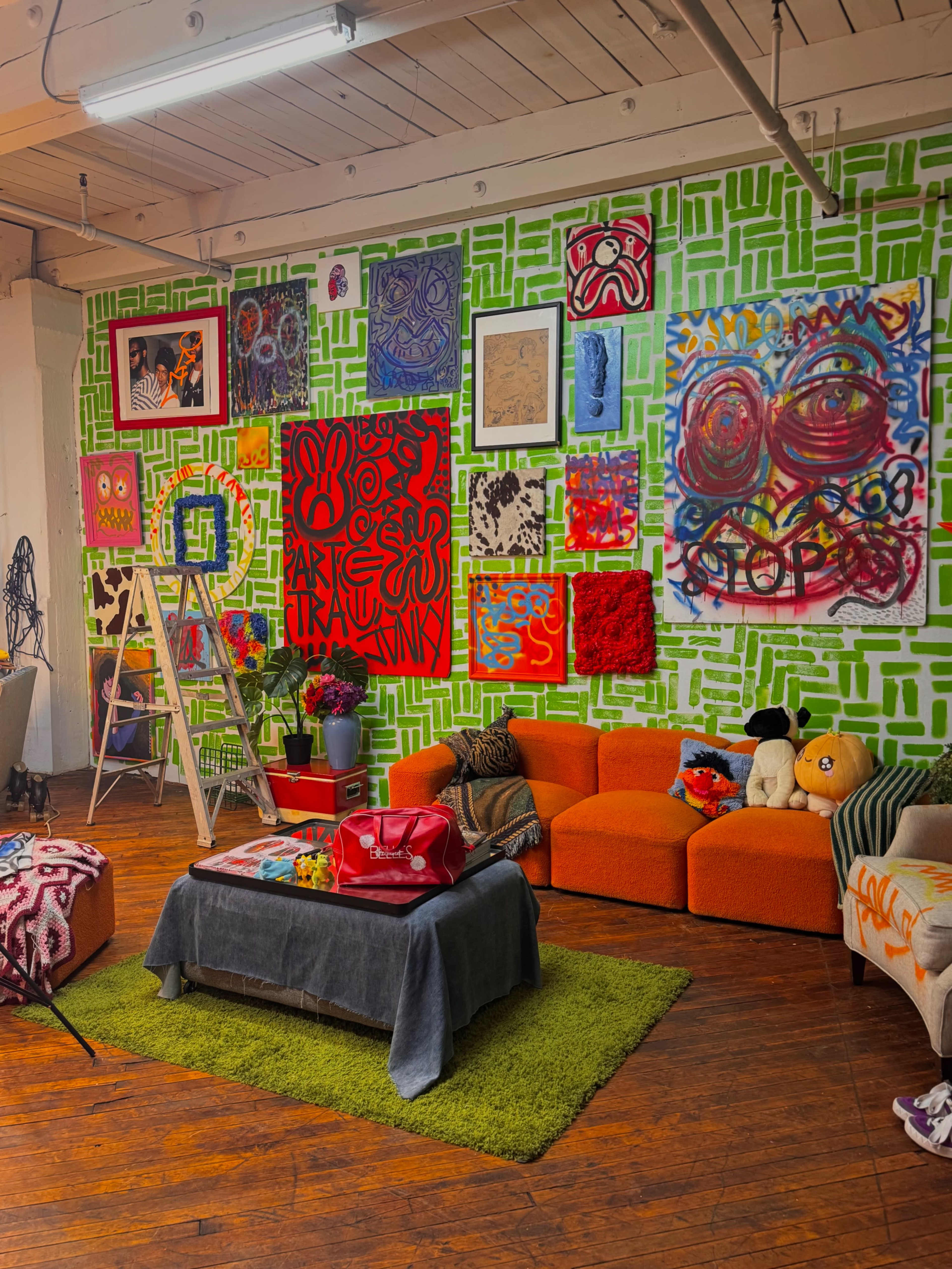 The image depicts a vibrant art studio with a green and white patterned wall adorned with various colorful artworks, featuring a seating area with orange sofas and a coffee table.