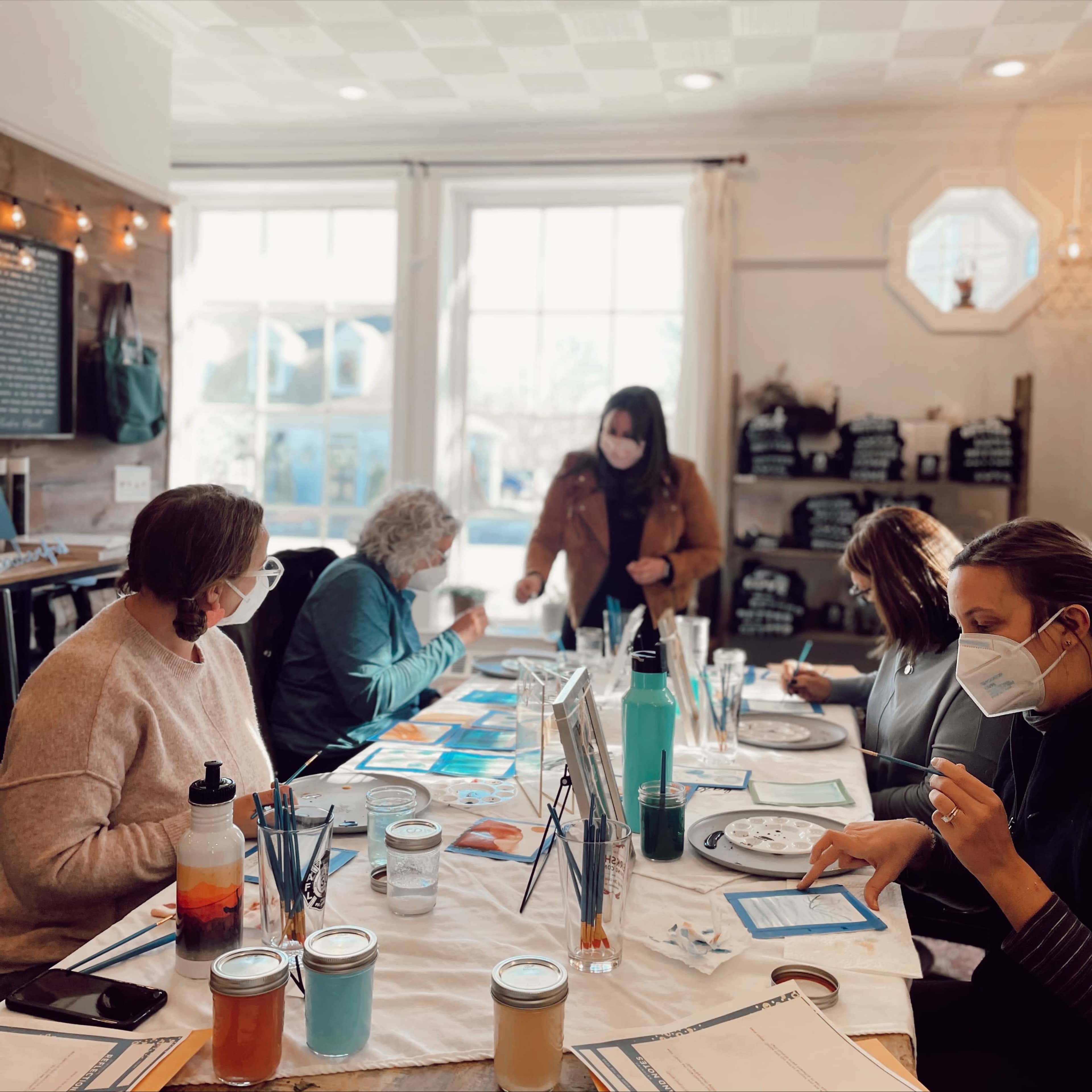 A group of adults sit around a table in a bright room, engaged in a painting activity while wearing masks.