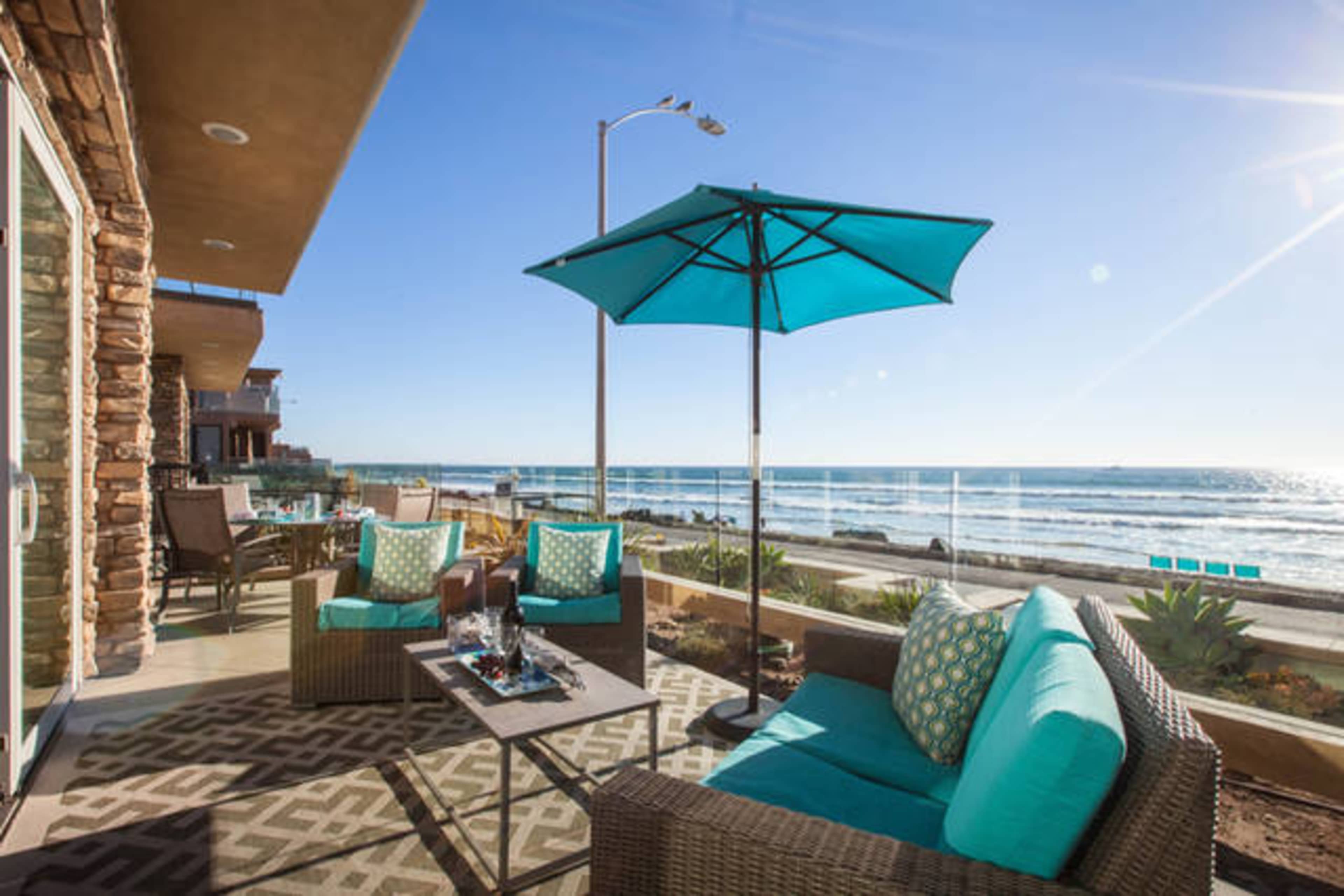 A patio area with turquoise umbrellas and chairs overlooks the ocean and waves under a clear blue sky.