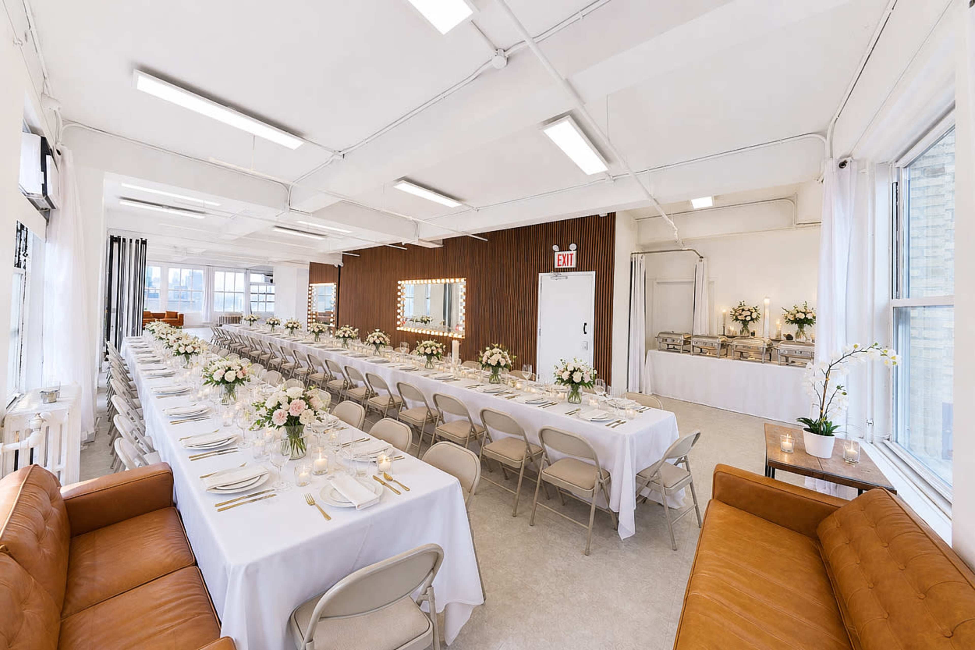 The image shows a spacious event room set with long tables covered in white tablecloths, arranged with floral centerpieces.