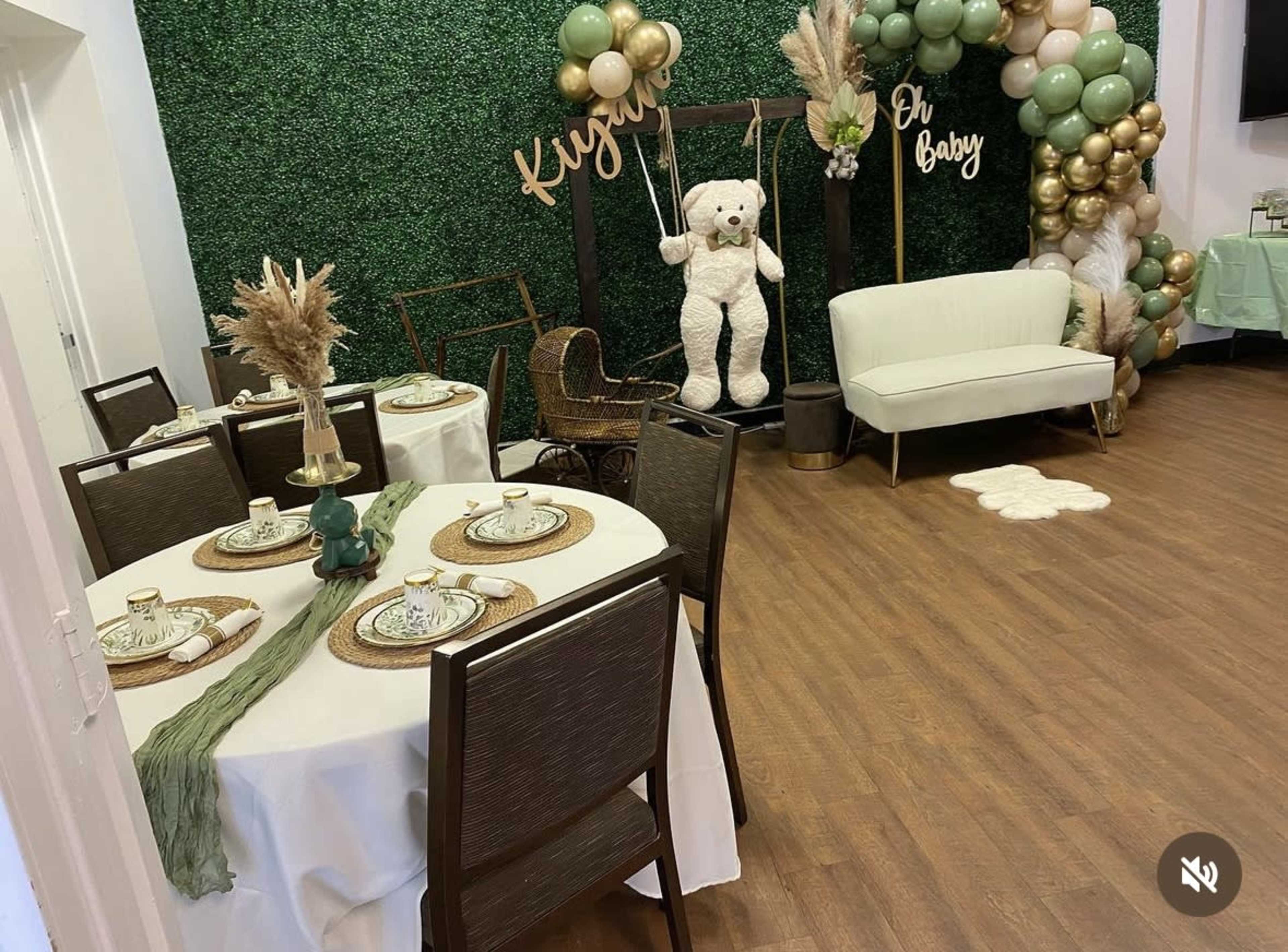 The image shows a decorated room for a baby shower, featuring green foliage on the wall, tables set with dishes, and a large teddy bear near a seating area adorned with balloons and decorative elements.