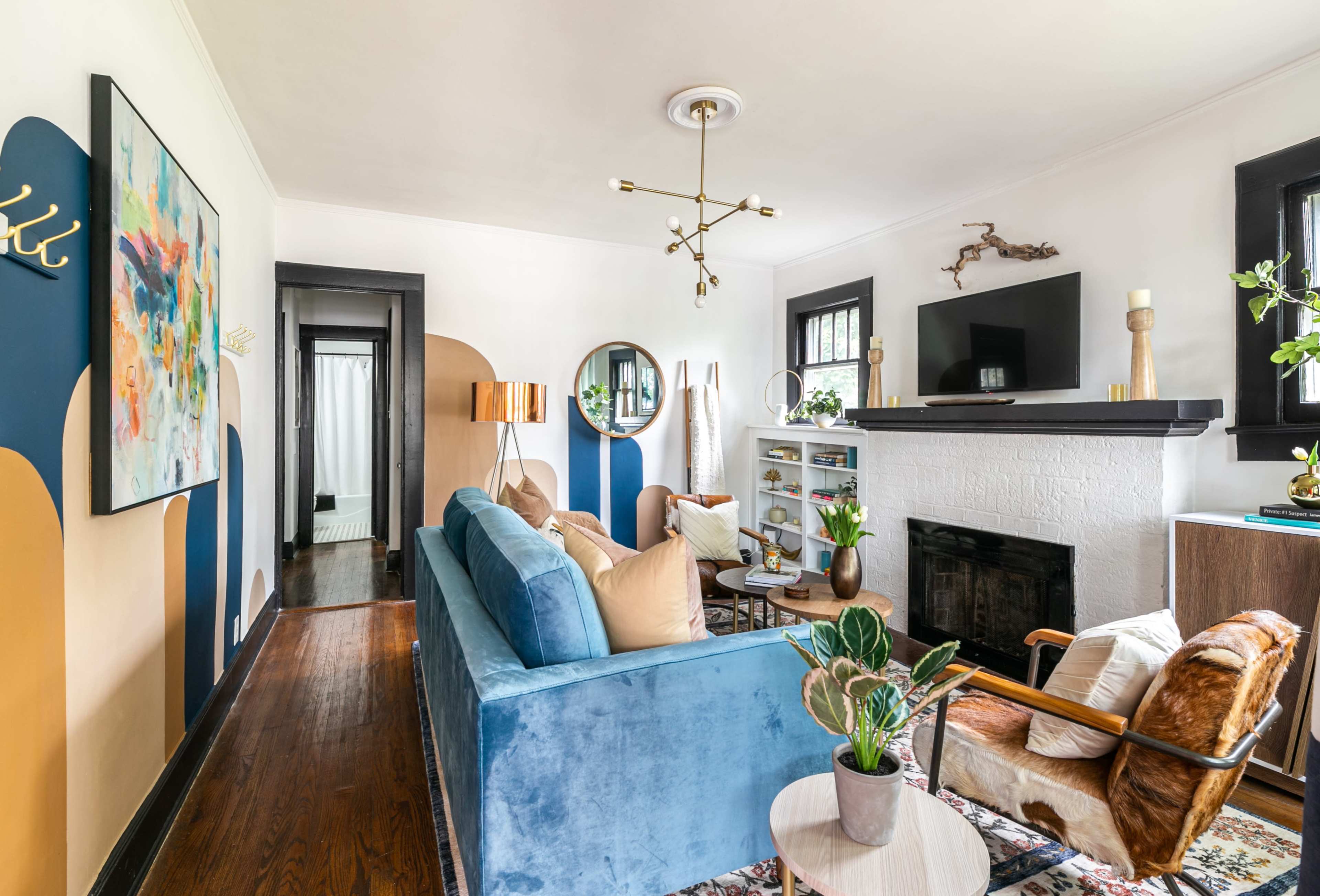 A living room with a blue sofa, an armchair, a coffee table, a bookshelf, and a fireplace, all accented by a colorful wall mural and modern lighting.