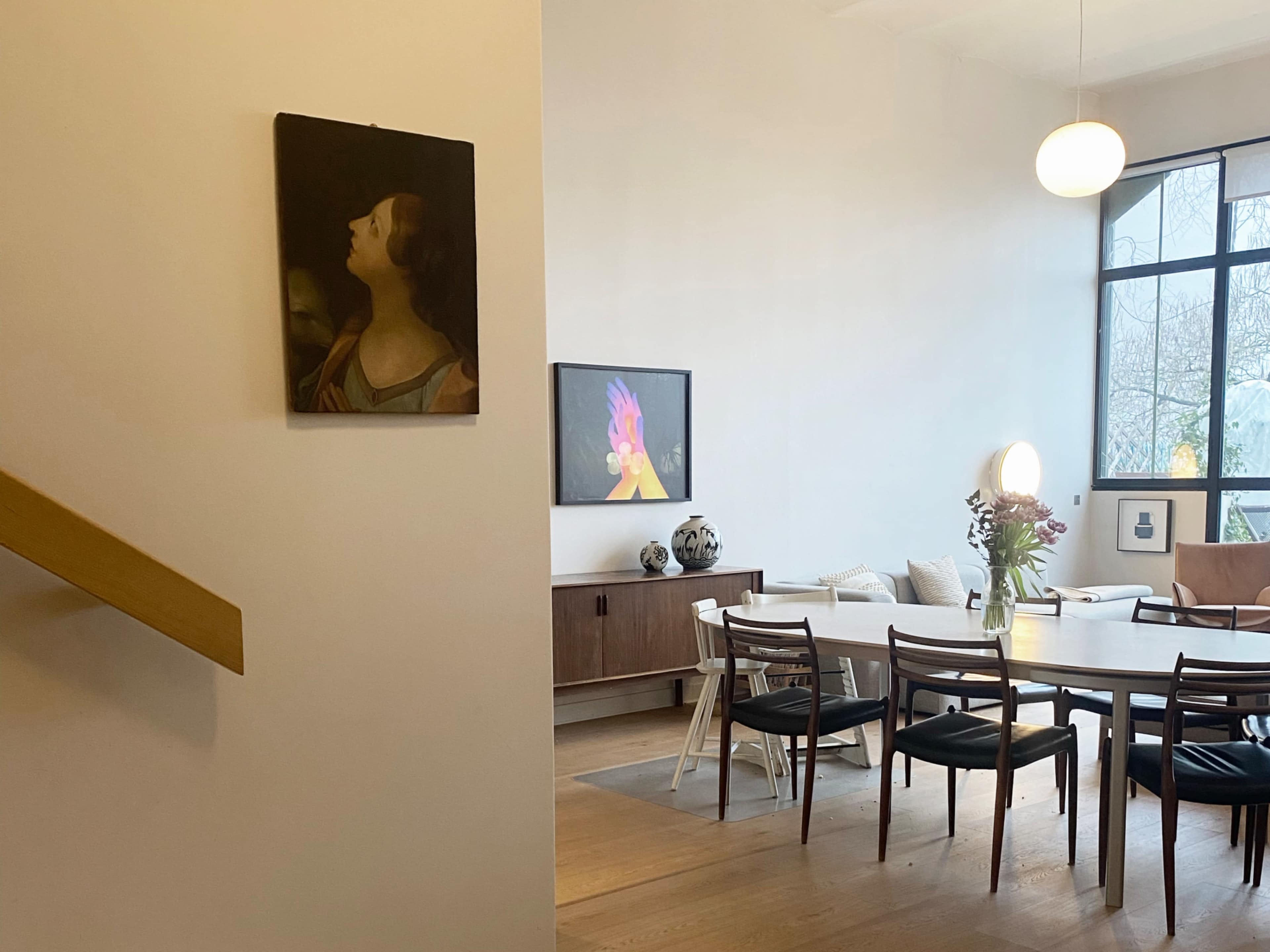 The image shows a modern dining area with a table surrounded by chairs, a painting on the wall, and a television mounted nearby.