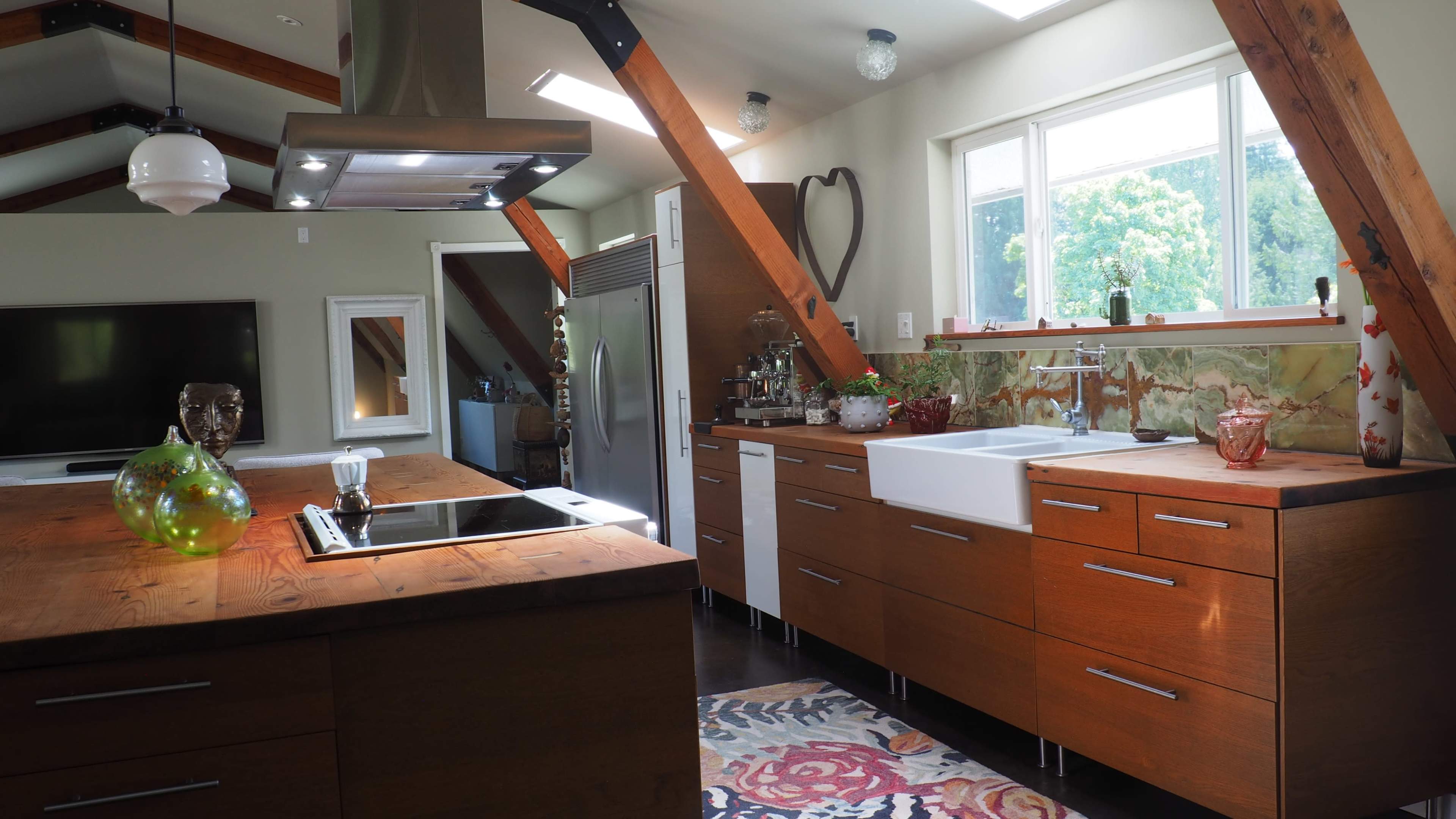 The image shows a spacious modern kitchen featuring wooden cabinets, a large sink, and an island with an integrated cooktop, framed by exposed wooden beams and bright natural light.