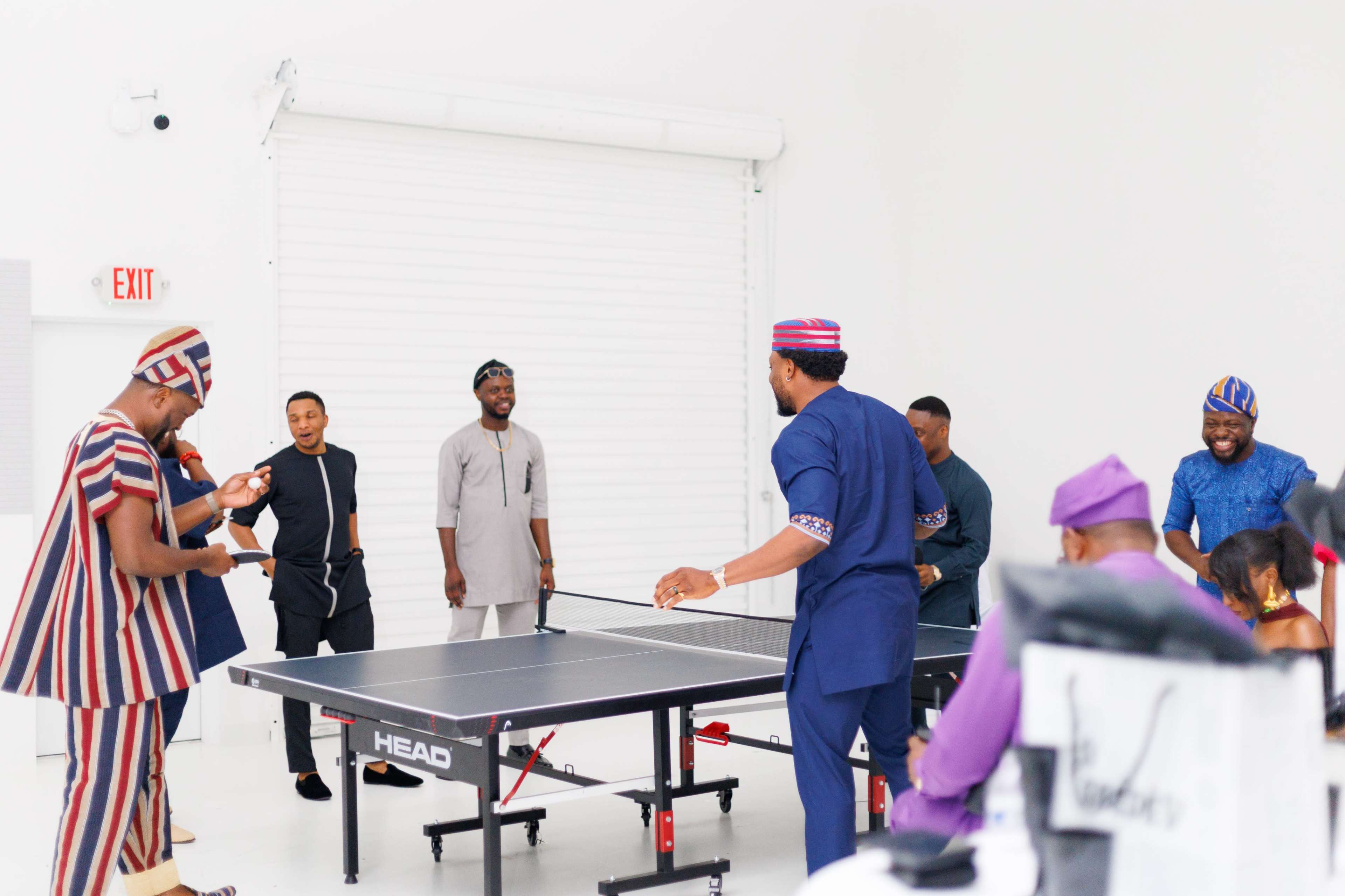 A group of men is playing table tennis in a brightly lit, minimalistic room with a roll-up door.