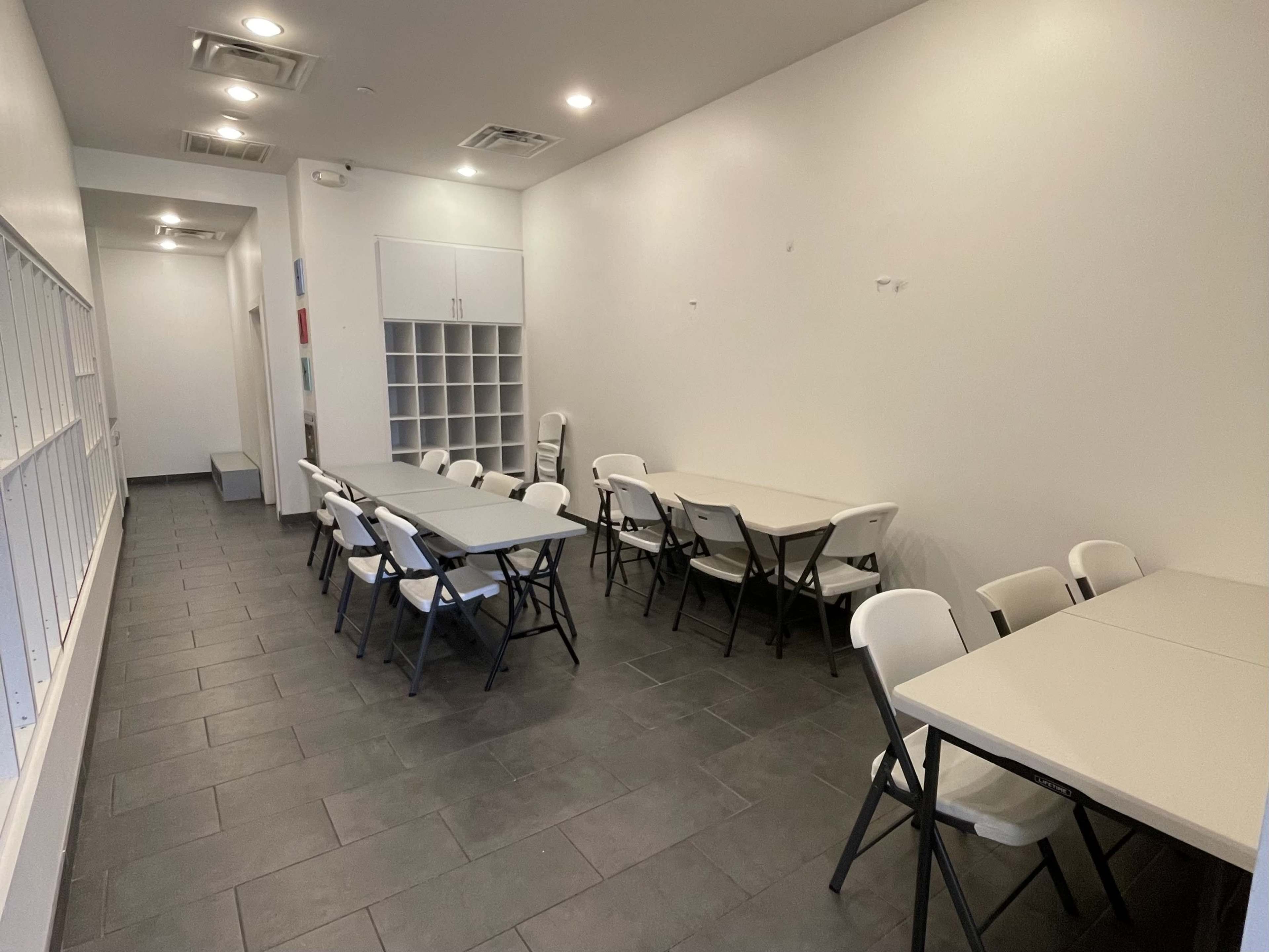 The image shows a bright, empty room with several folding tables and chairs arranged neatly on a tiled floor.