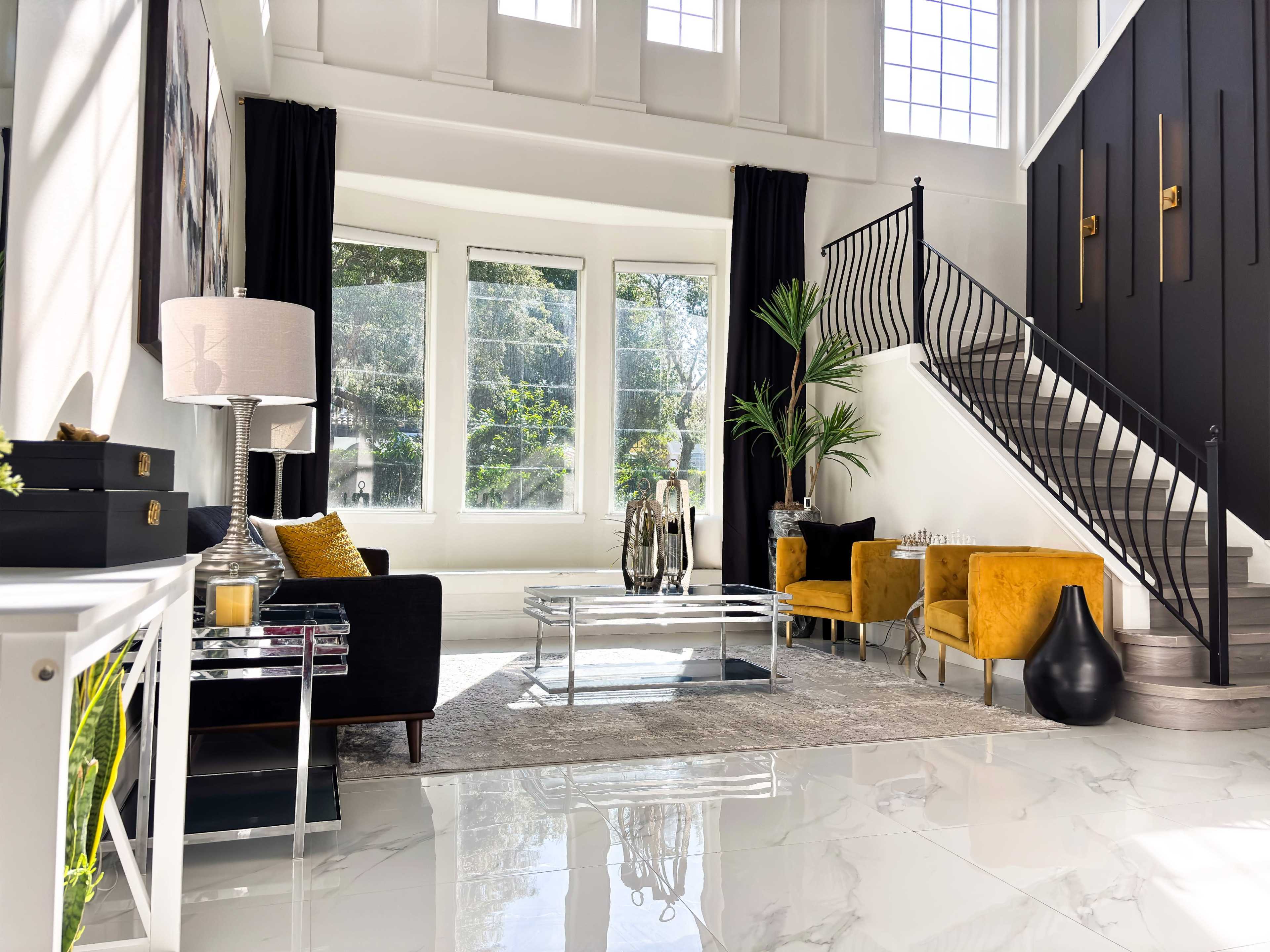The image shows a stylish living room featuring a black sofa, yellow chairs, a glass coffee table, and a staircase with ornate railings under bright natural light from large windows.