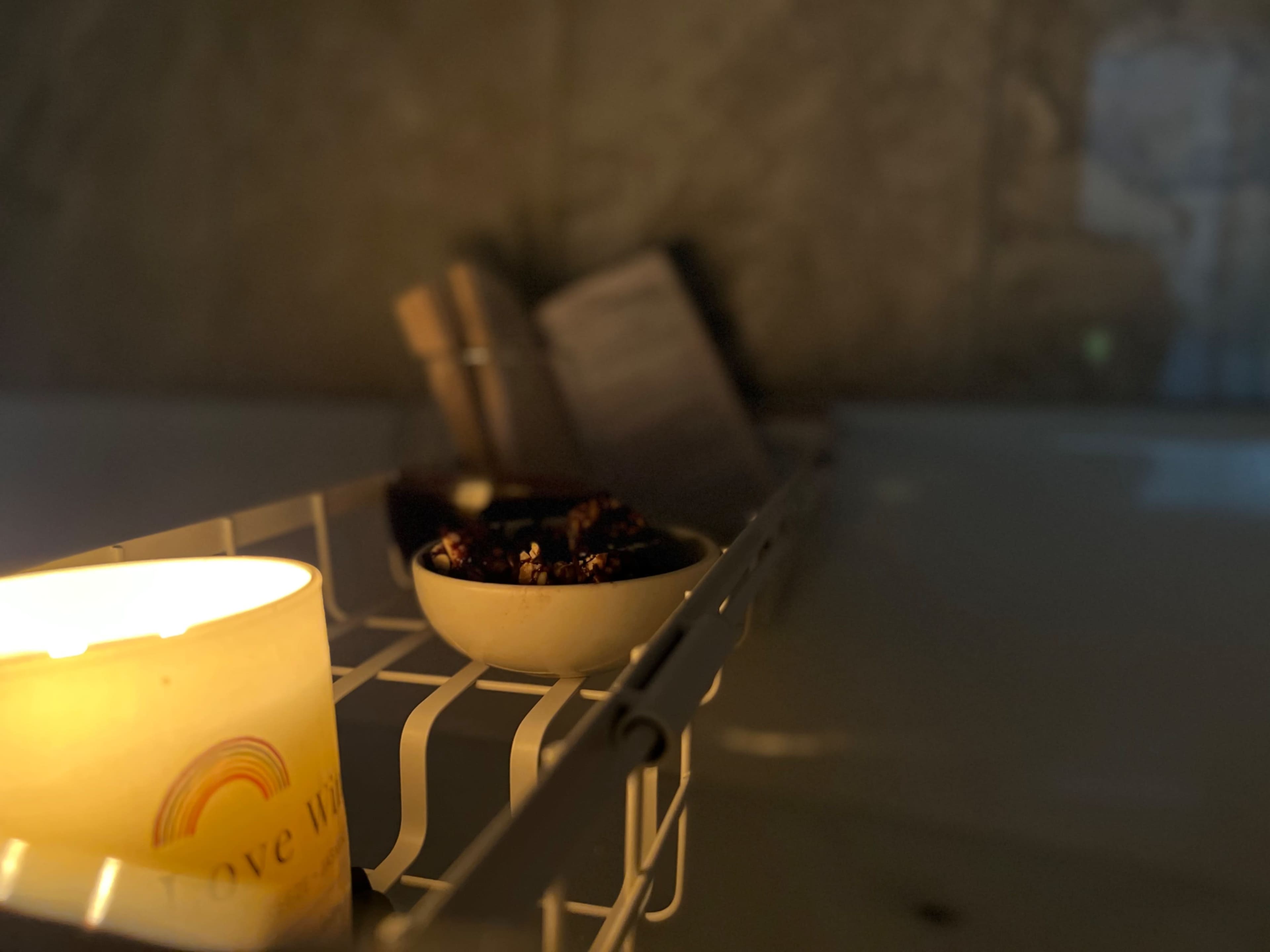 A lit candle and a small bowl of dark food items are set on a white rack near a bathtub.
