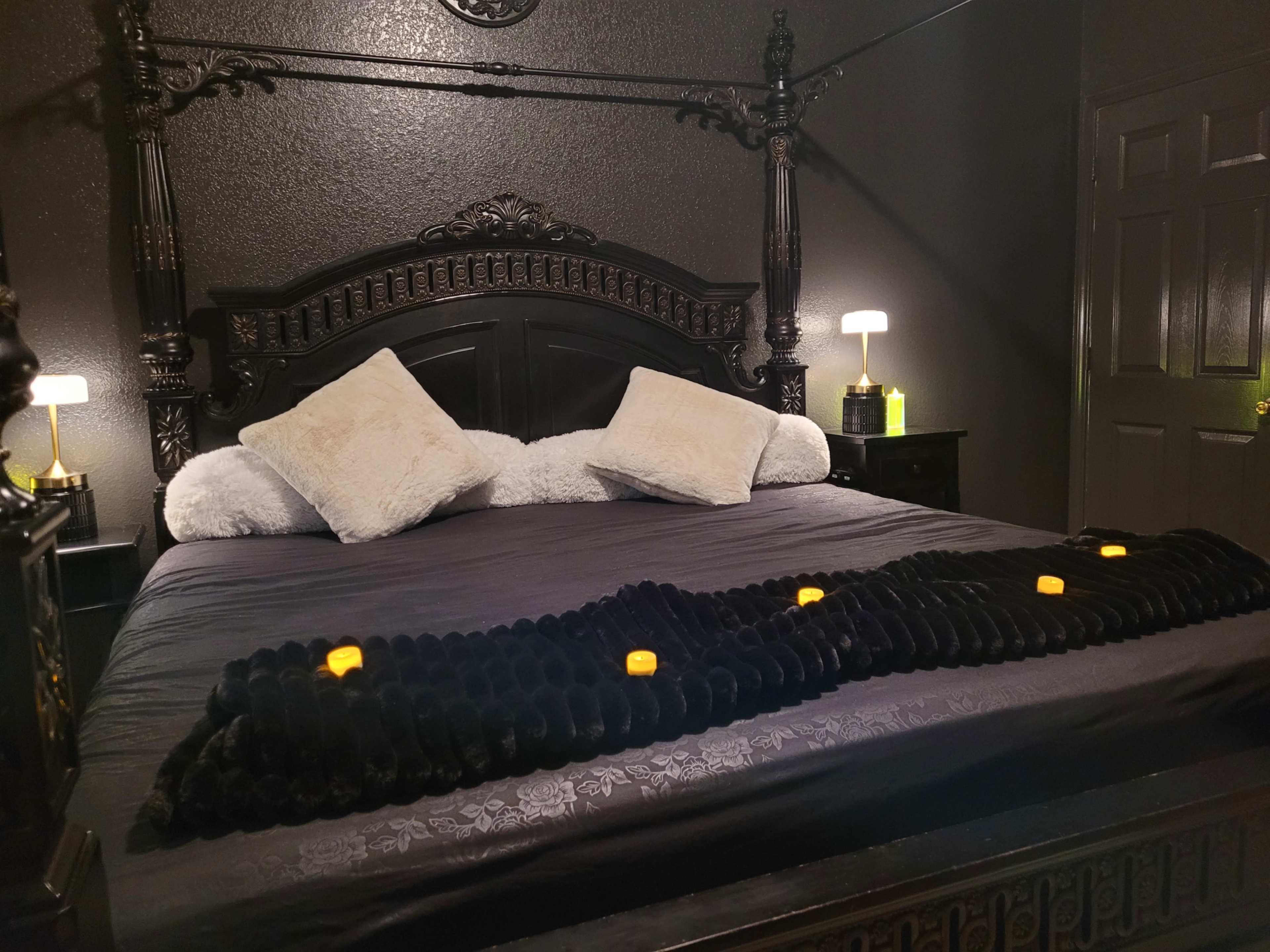 A large black bed with decorative posts features a dark bedding set, two white pillows, and a textured black throw blanket adorned with small candles.