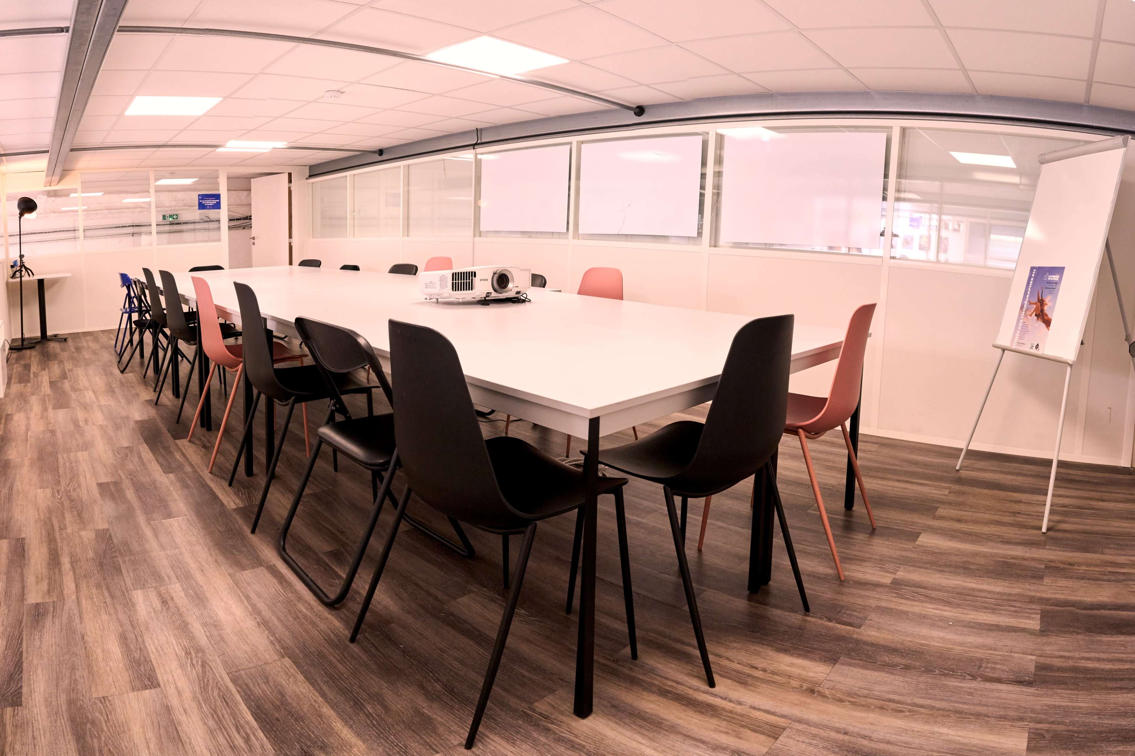 A spacious conference room features a long white table surrounded by black and pink chairs, with a projector on the table and a flip chart on the side.