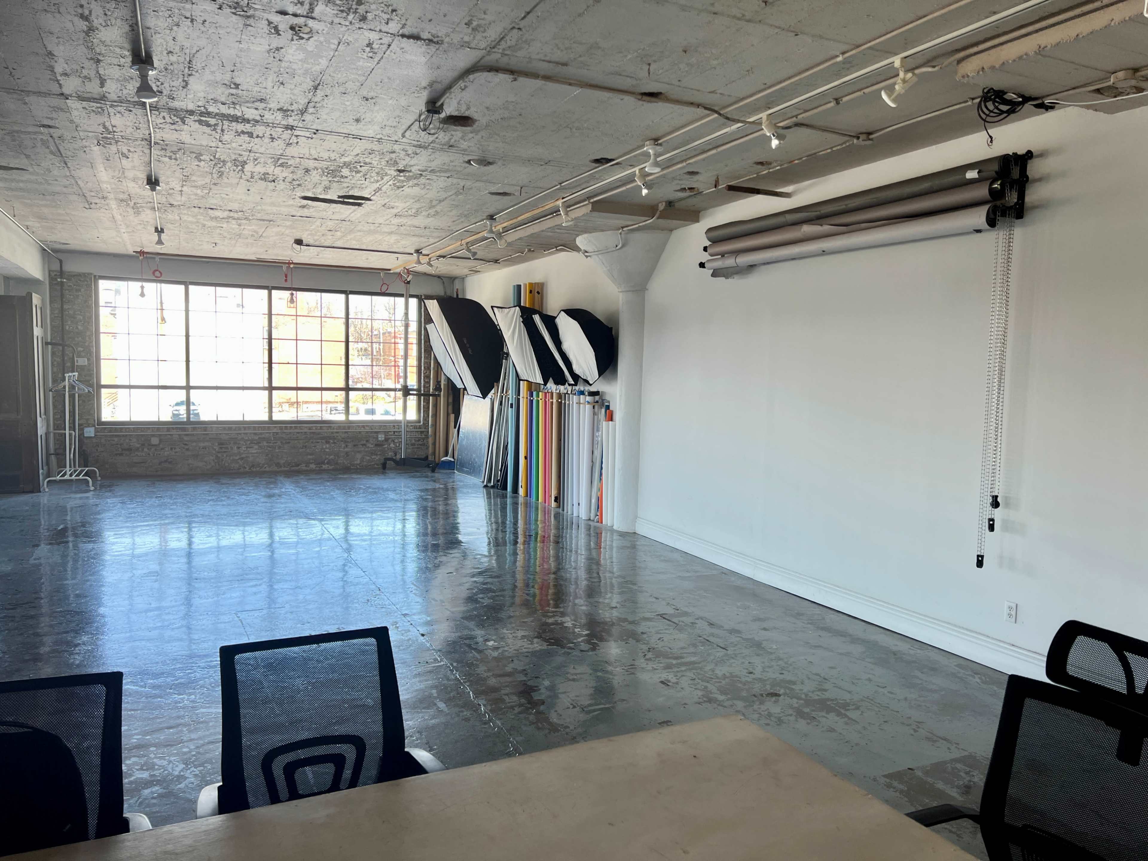 The image shows a spacious, well-lit studio with concrete floors, large windows, and various photography backdrops hanging on the wall.