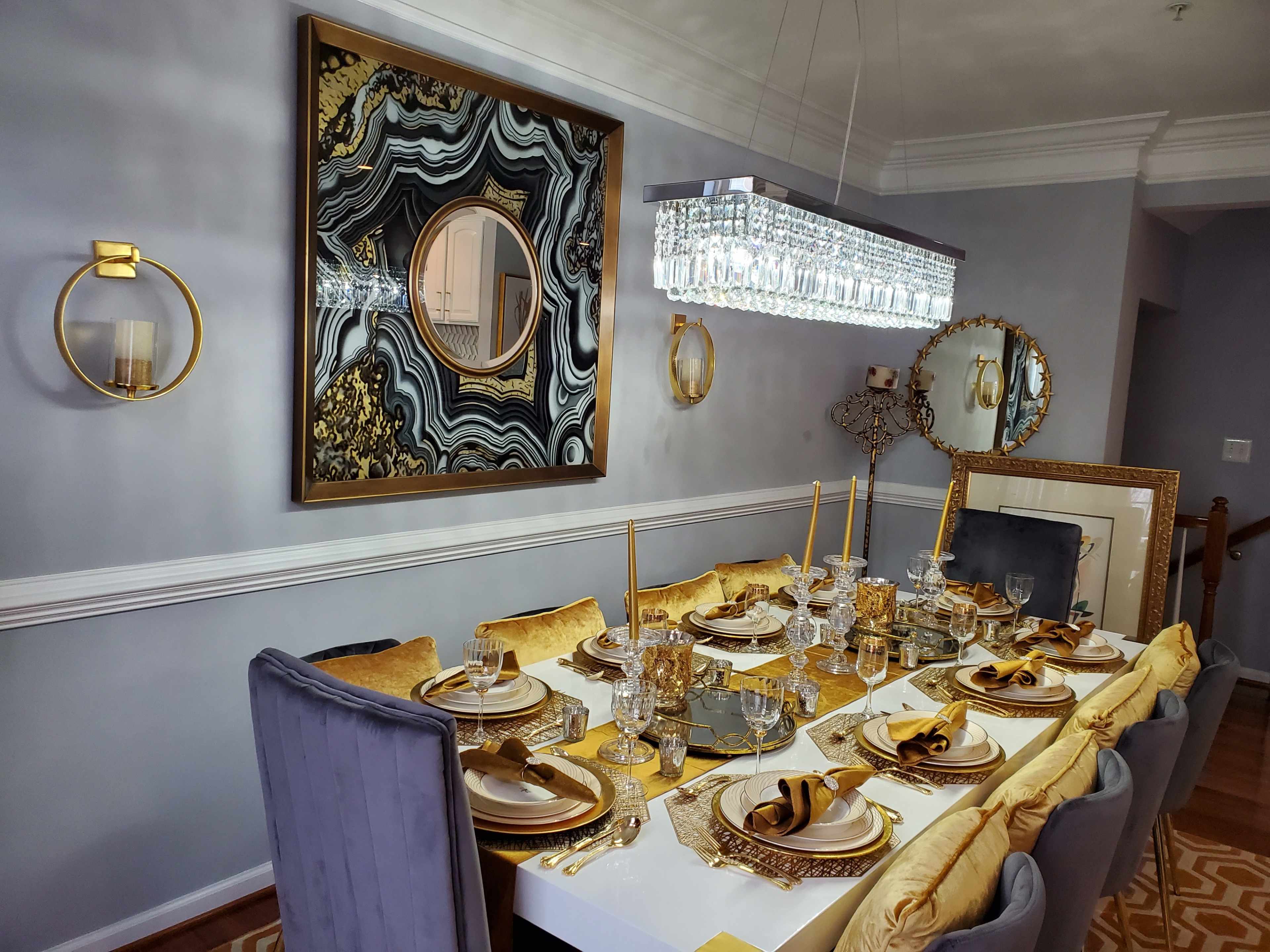 The dining room features an elegantly set table with gold accents, surrounded by plush chairs beneath a chandelier, all set against a backdrop of stylish wall decor.