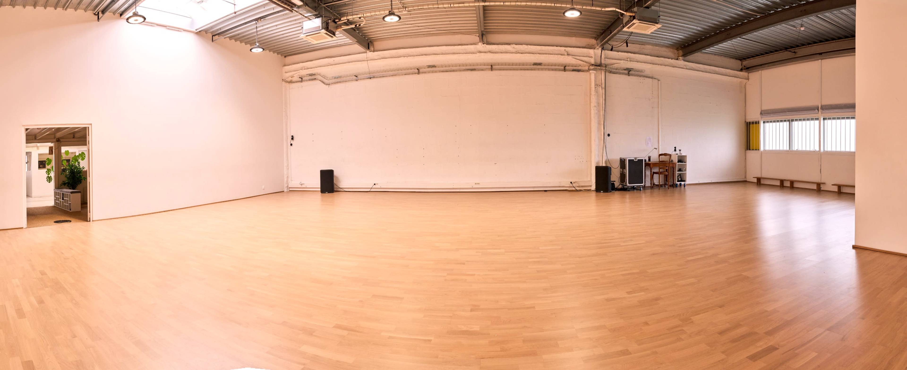 The image shows a spacious, empty room with a smooth wooden floor, white walls, and a large window, along with some furniture placed against one wall.