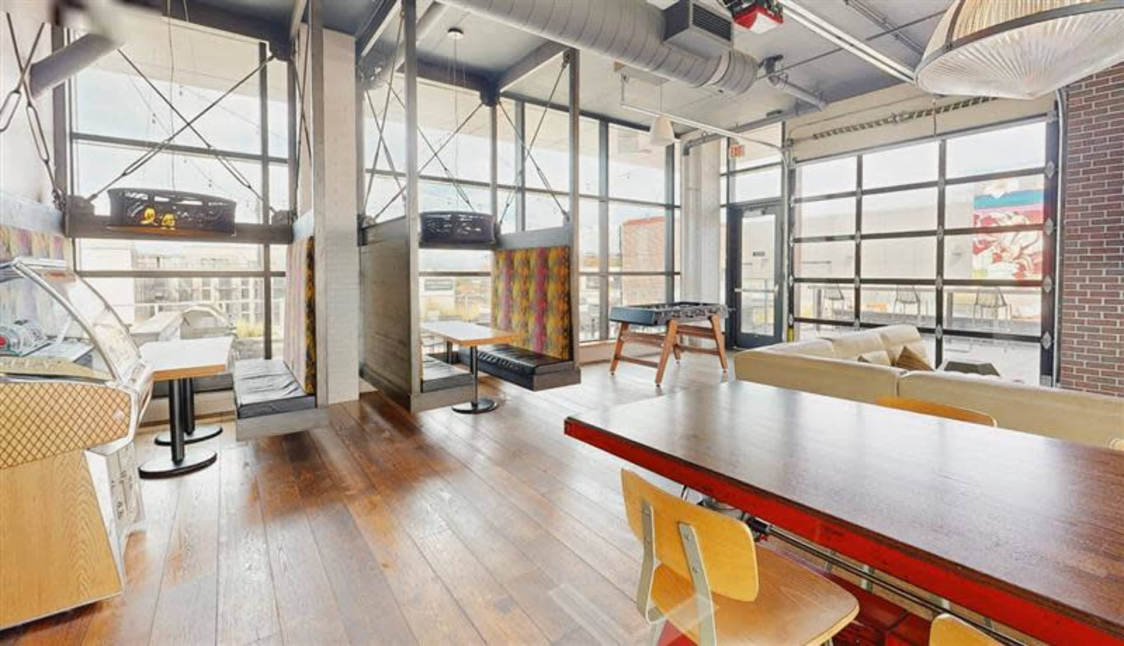 The image shows a modern, open space with large windows, wooden flooring, and various seating areas, including a dining table and lounge couches.