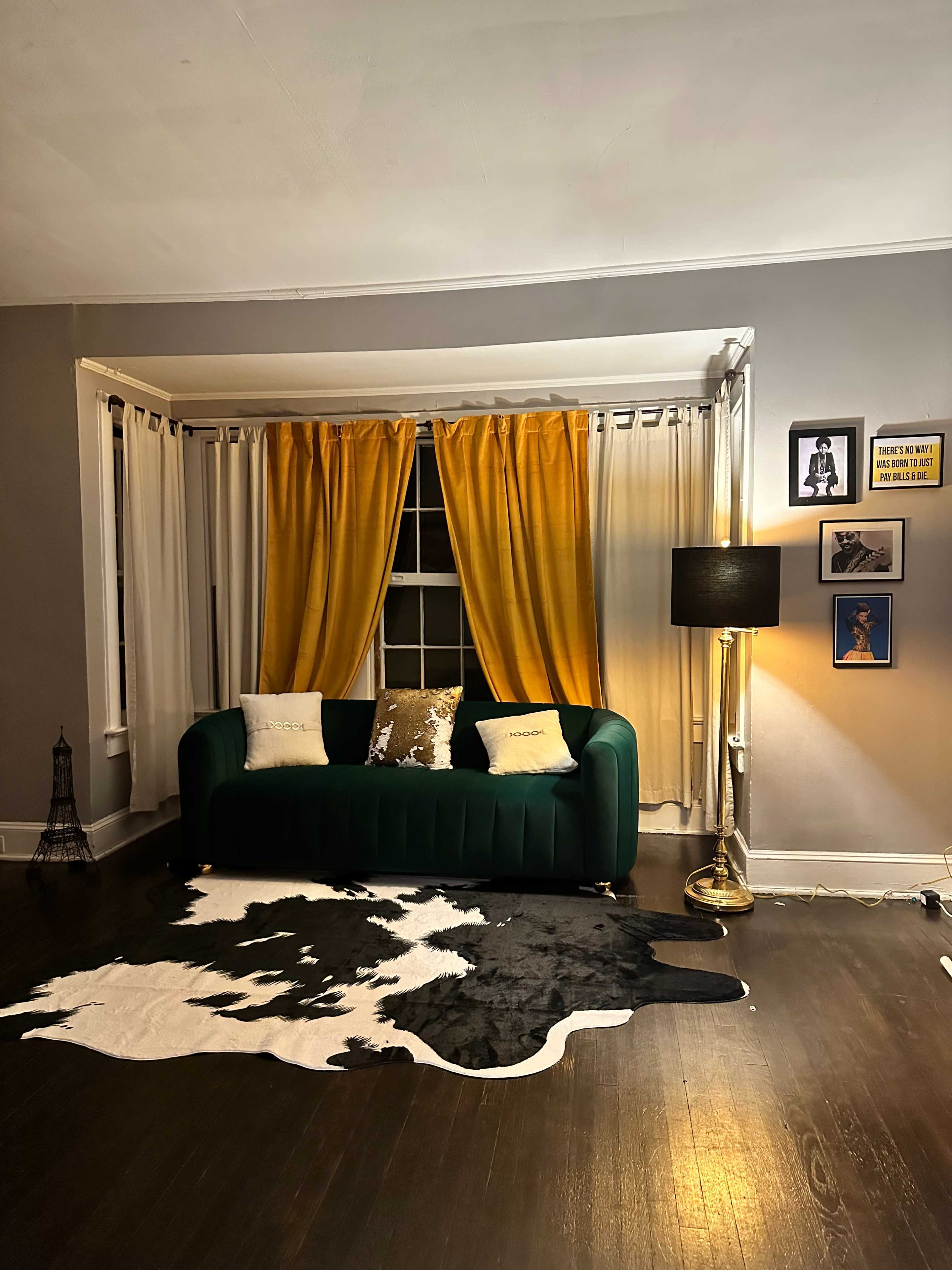 A living room with a green sofa, yellow curtains, a black and white cowhide rug, and a standing lamp beside framed artwork on the wall.