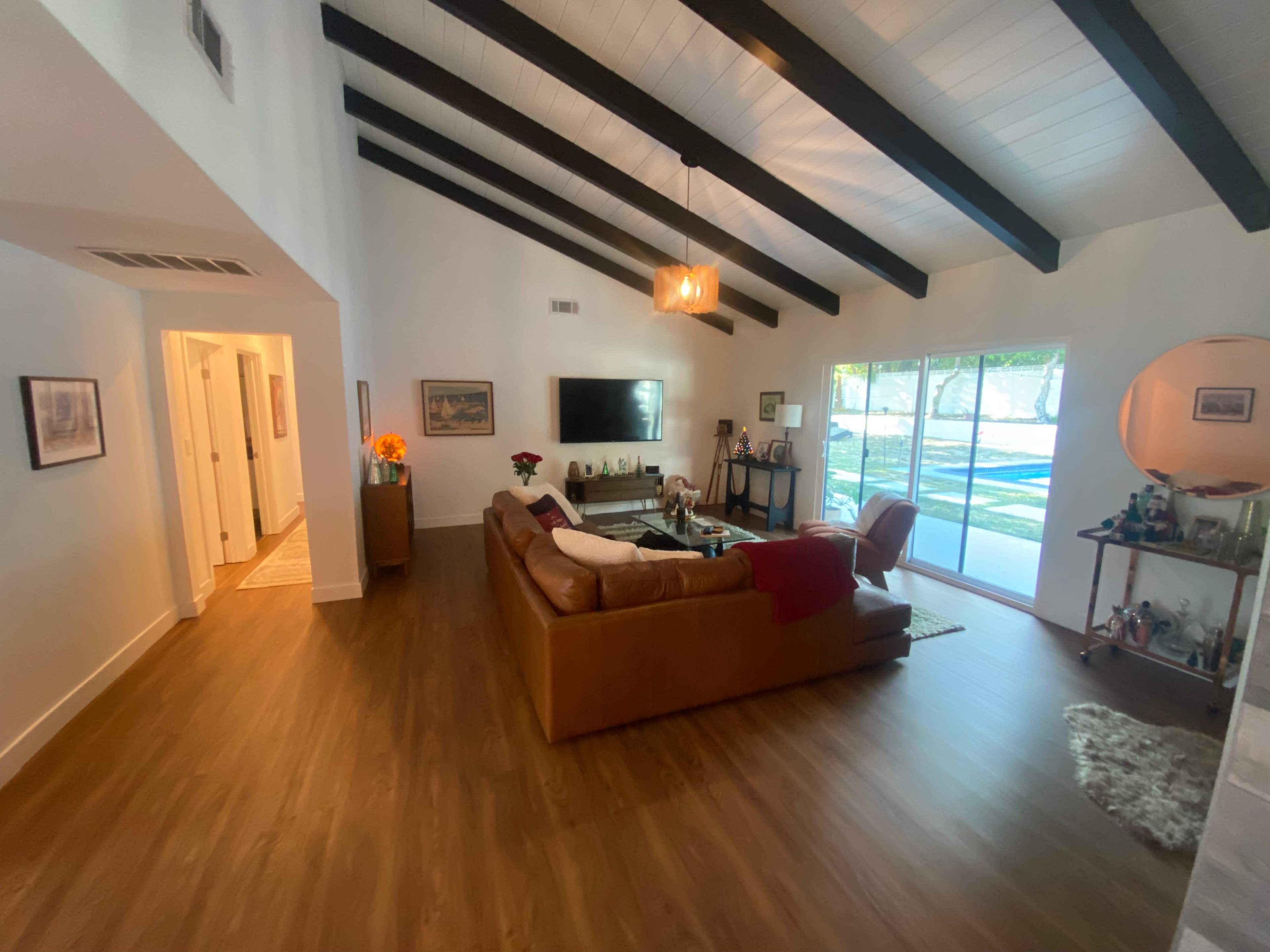 A spacious living room features a brown leather couch, a television on the wall, and large windows providing natural light.