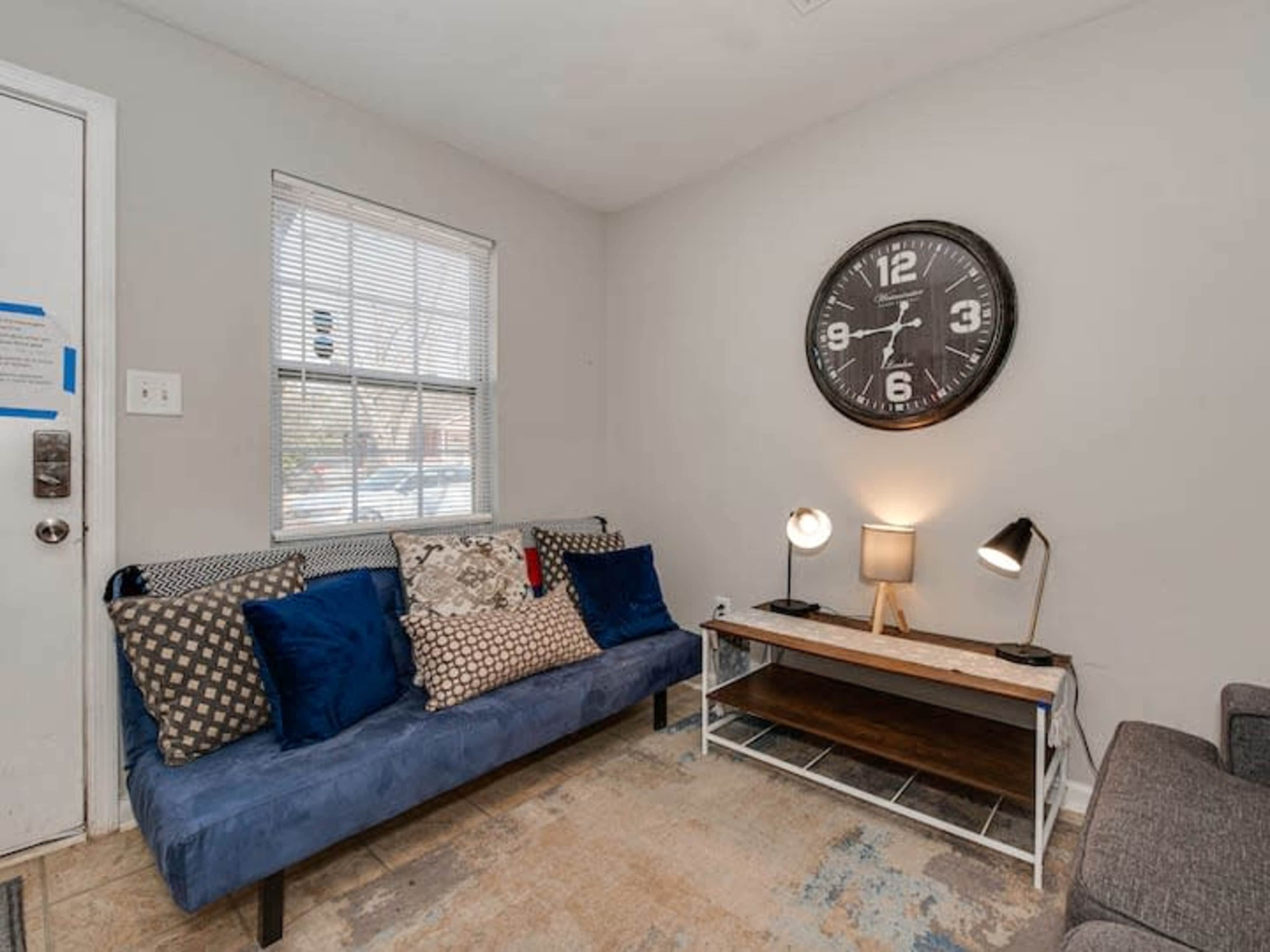 A small living area with a blue couch adorned with pillows, a wooden coffee table, two lamps, and a wall clock.