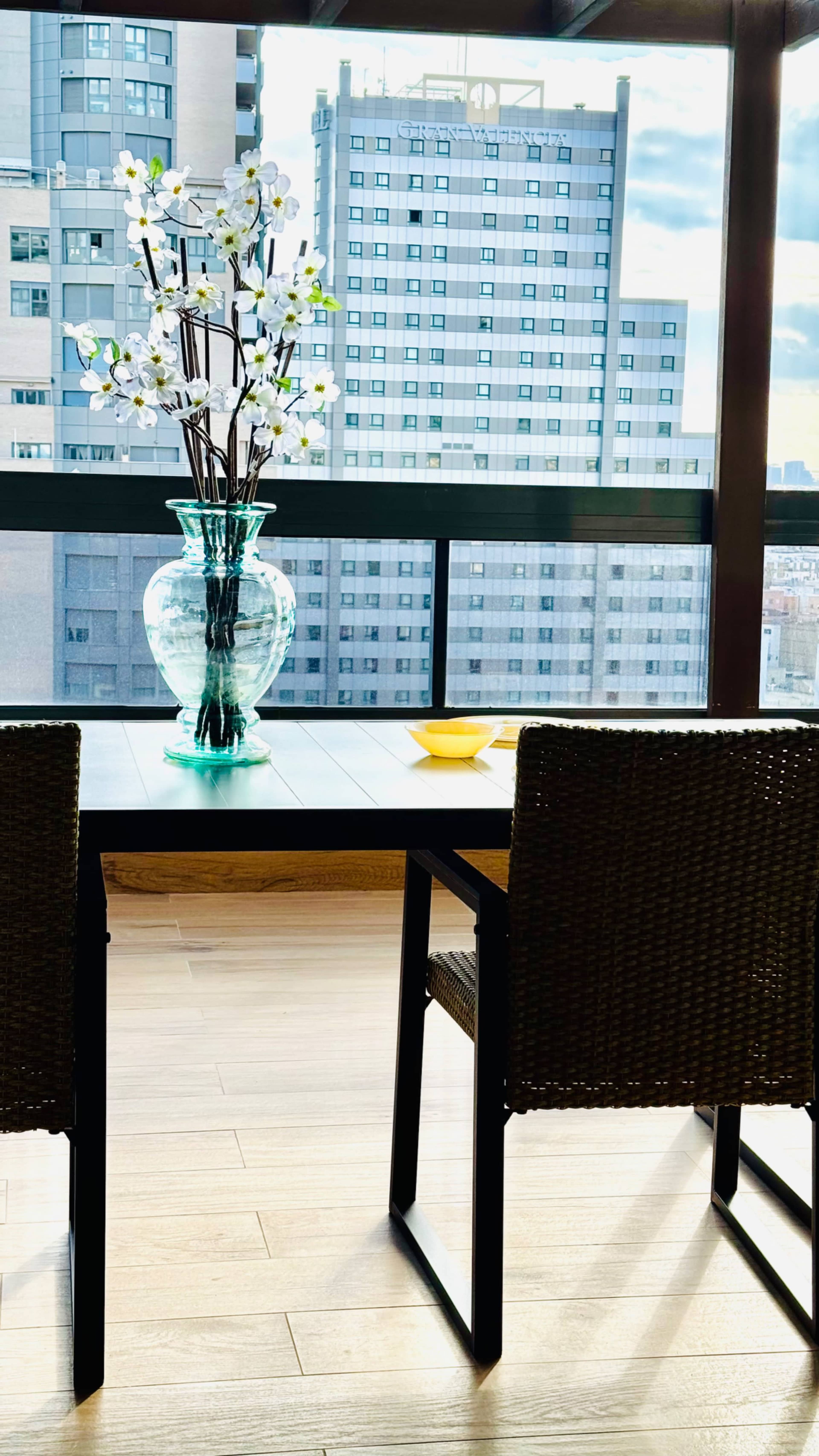 A glass vase filled with artificial flowers sits on a dining table, overlooking a city skyline through a large window.