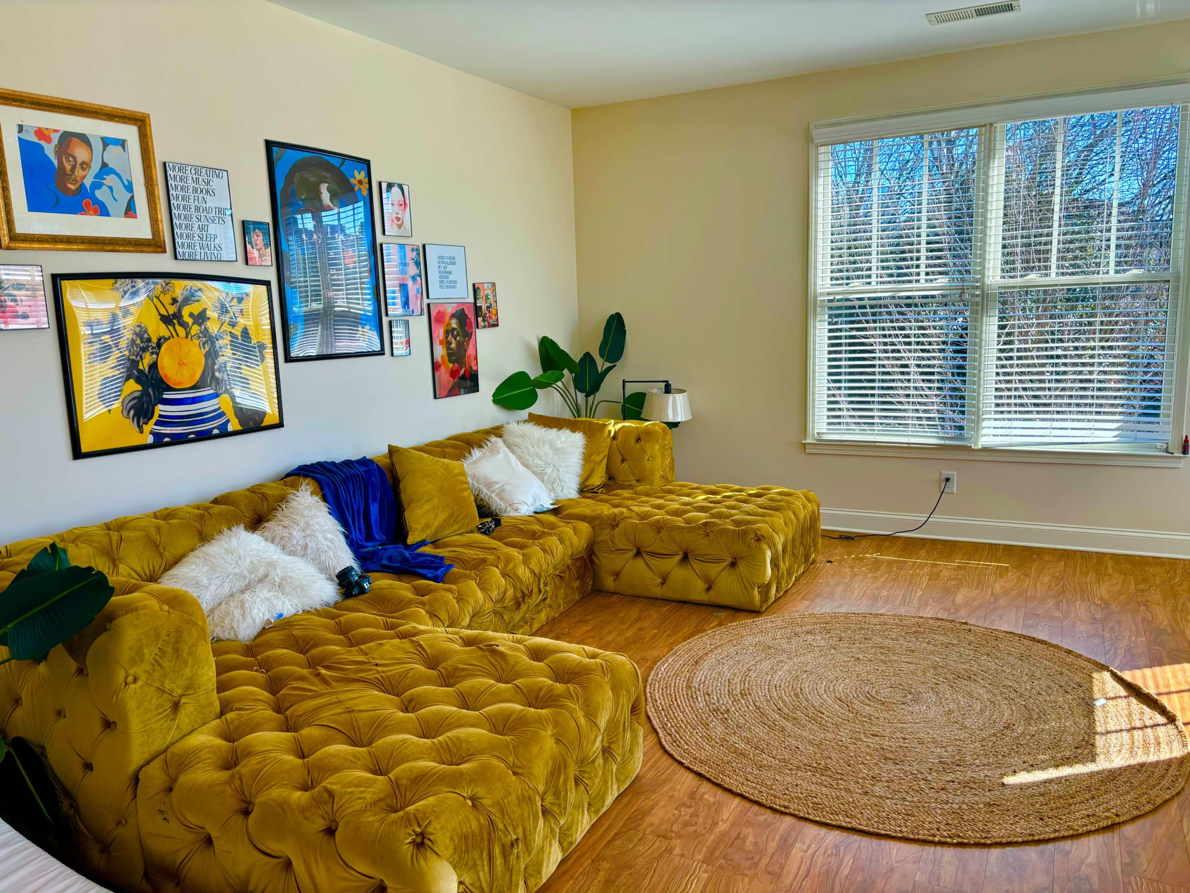 The scene features a bright living room with a yellow tufted sectional sofa, round jute rug, and large windows allowing natural light.
