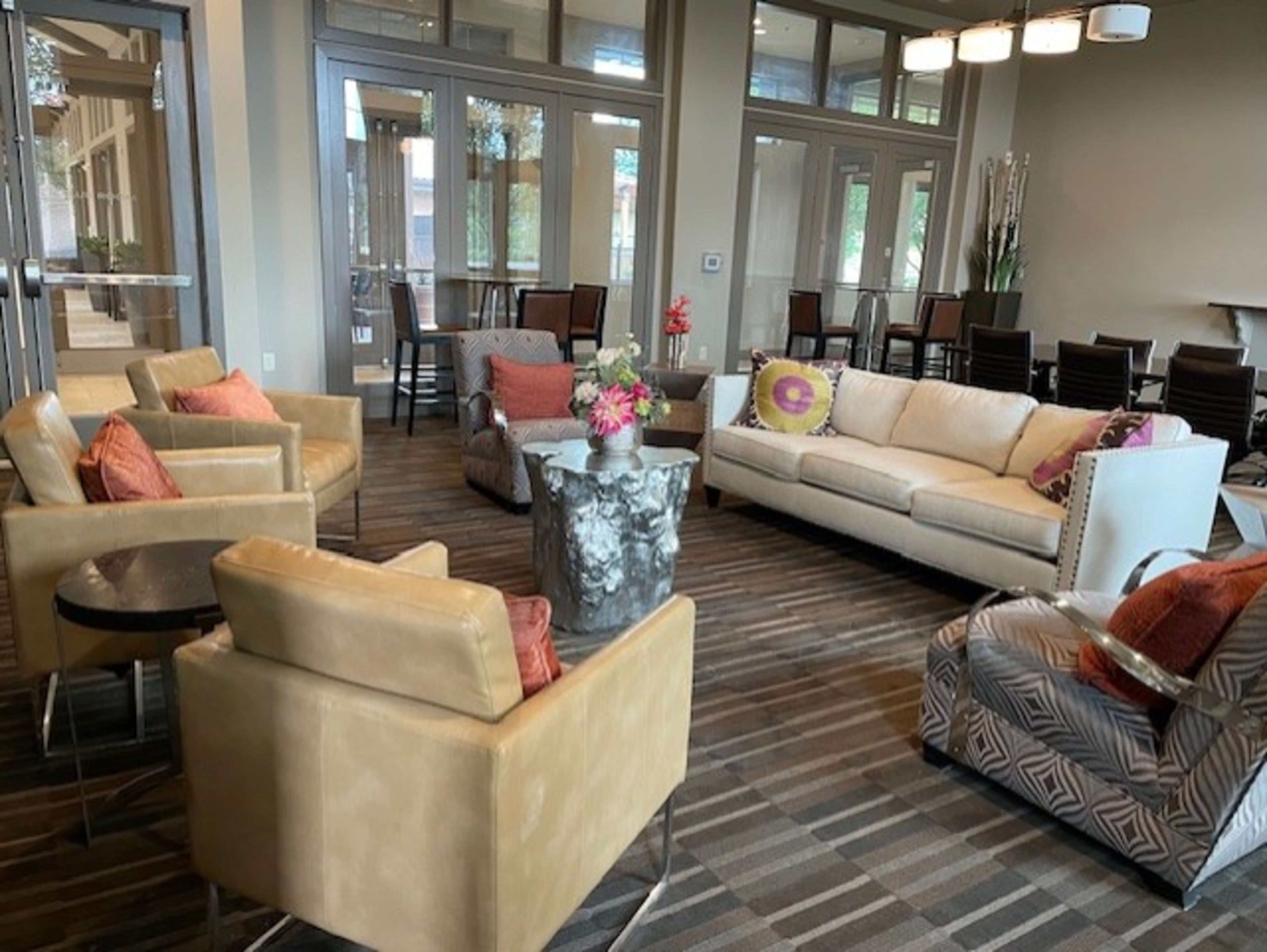 The image shows a modern lounge area with a sectional sofa, several armchairs, and a coffee table, surrounded by large windows and dining tables in the background.