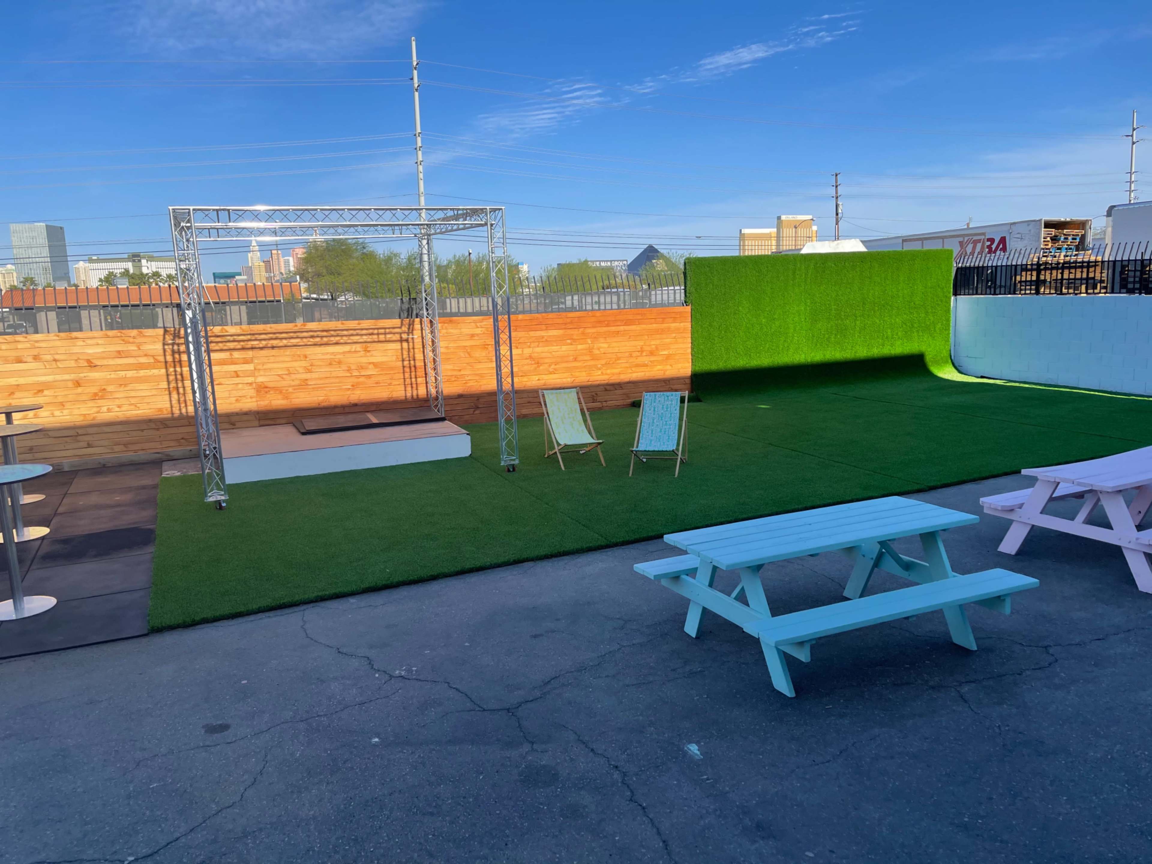 An outdoor venue featuring a grassy area with two light blue picnic tables, two chairs, and a performance stage framed by a green backdrop.