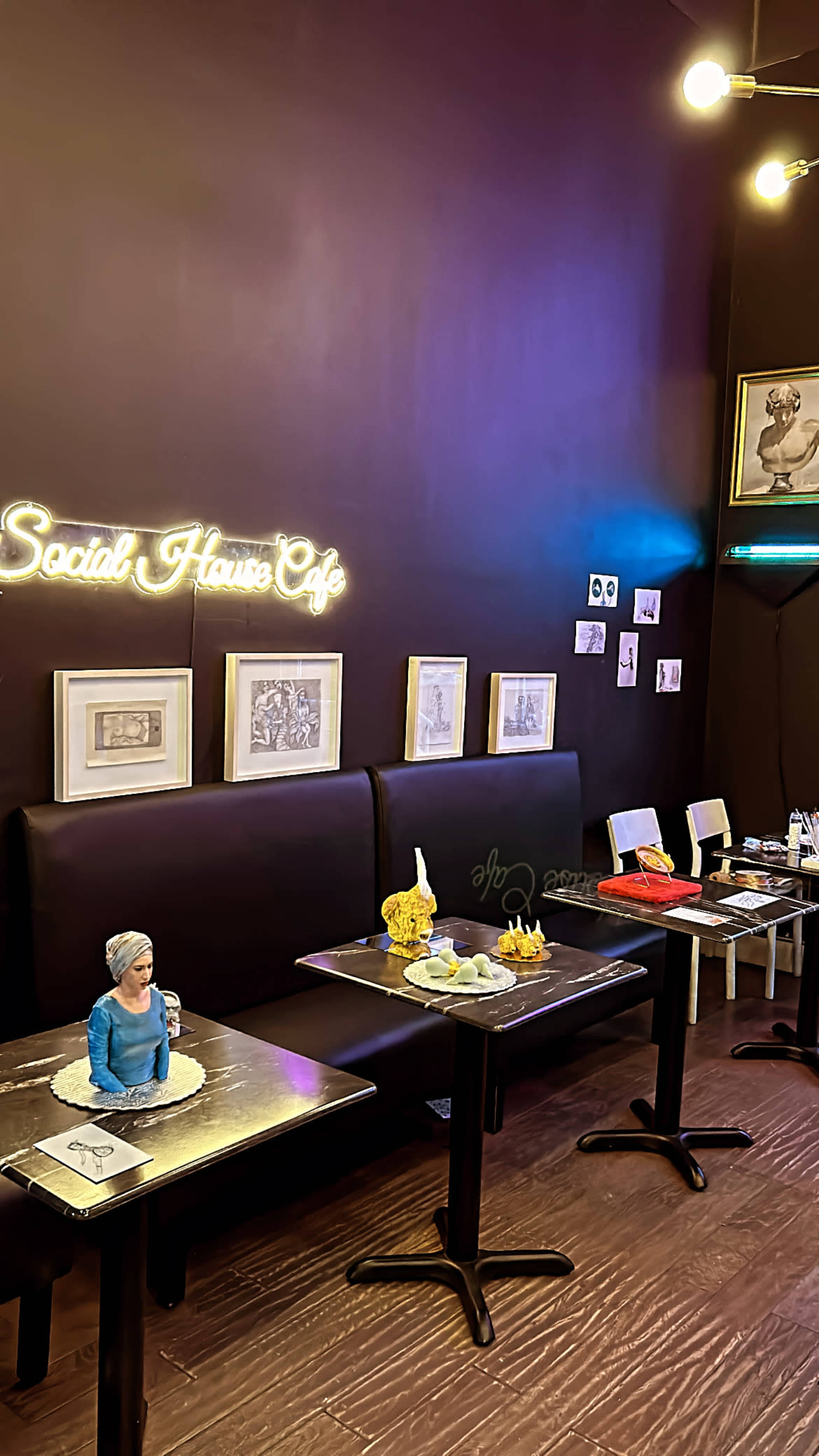 The image shows a café interior with a neon sign reading "Social House Café," featuring tables set with various decorative items and a sculpture on one table.