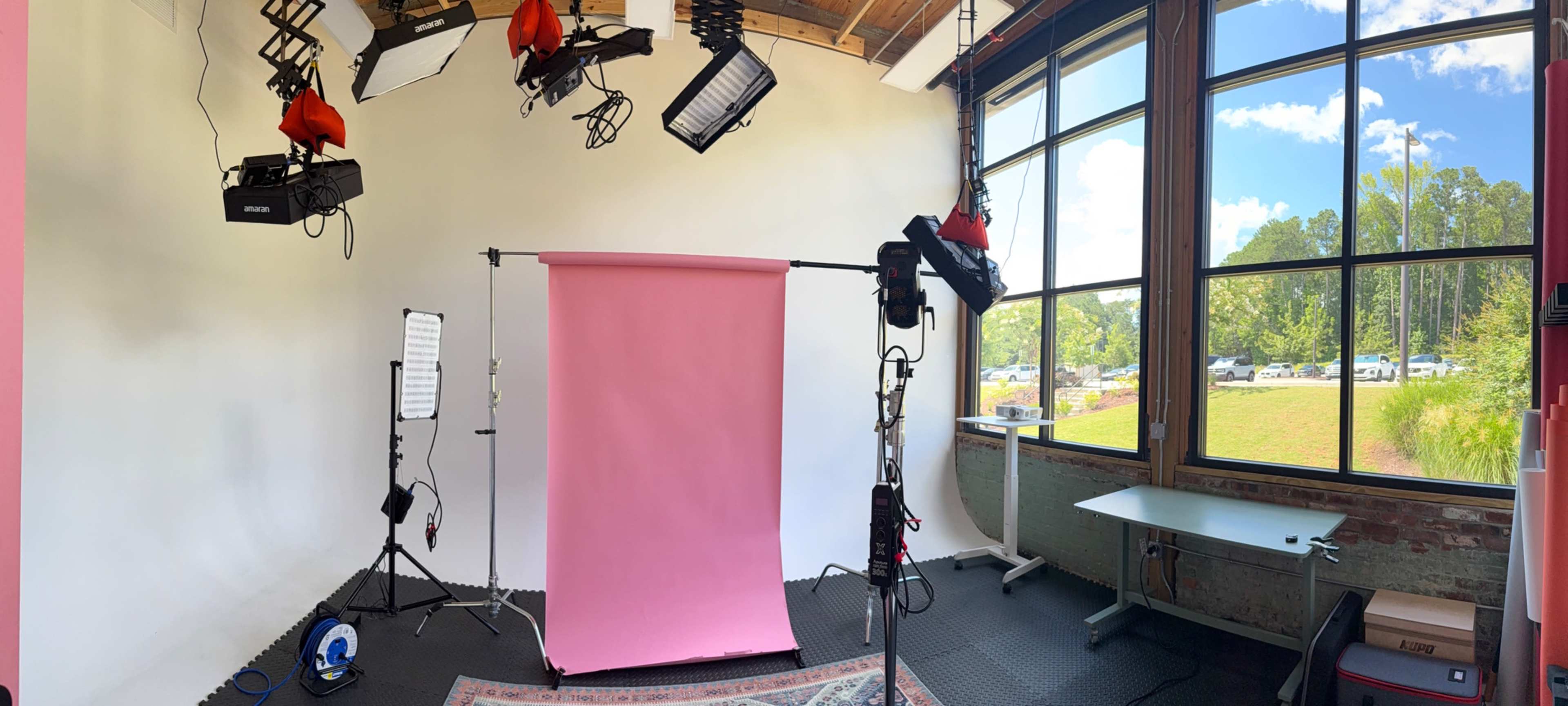 The image shows a photography studio equipped with various lighting setups, a pink backdrop, and large windows letting in natural light.
