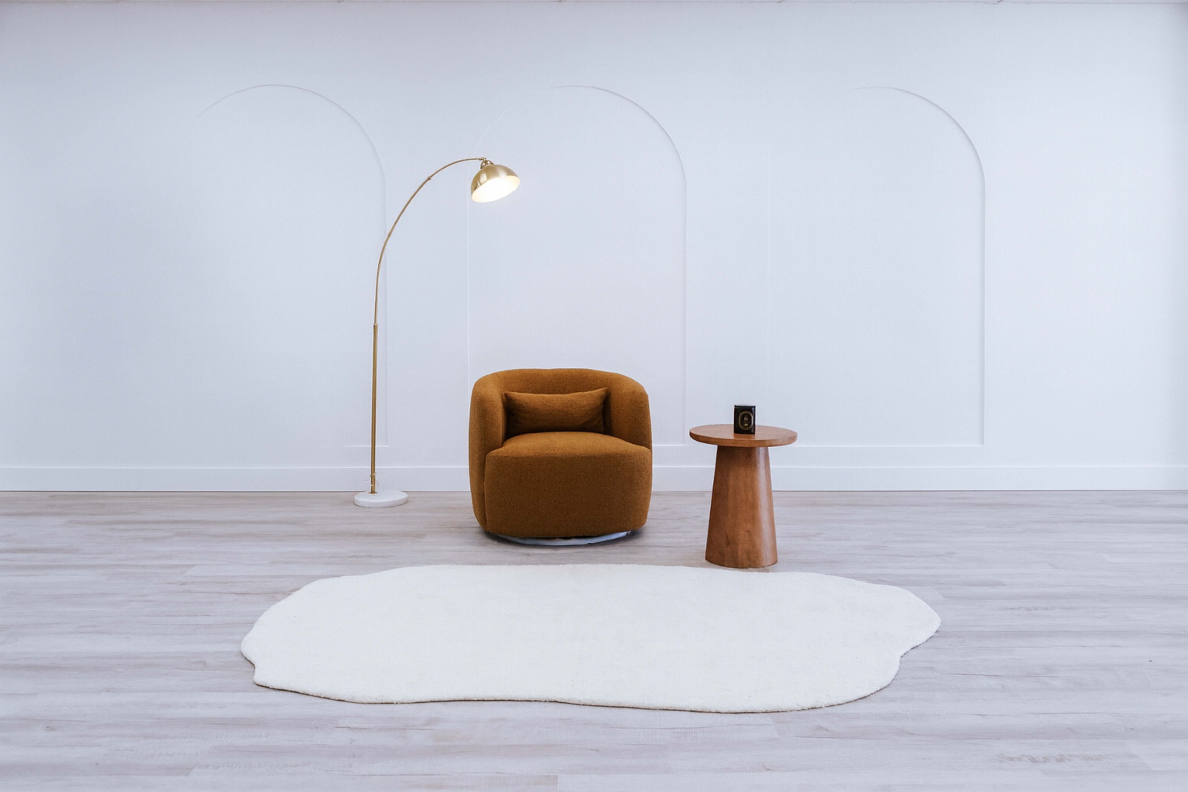A cozy, modern seating area features an orange armchair, a wooden side table, and a floor lamp, all set against a minimalistic white wall with arch details.