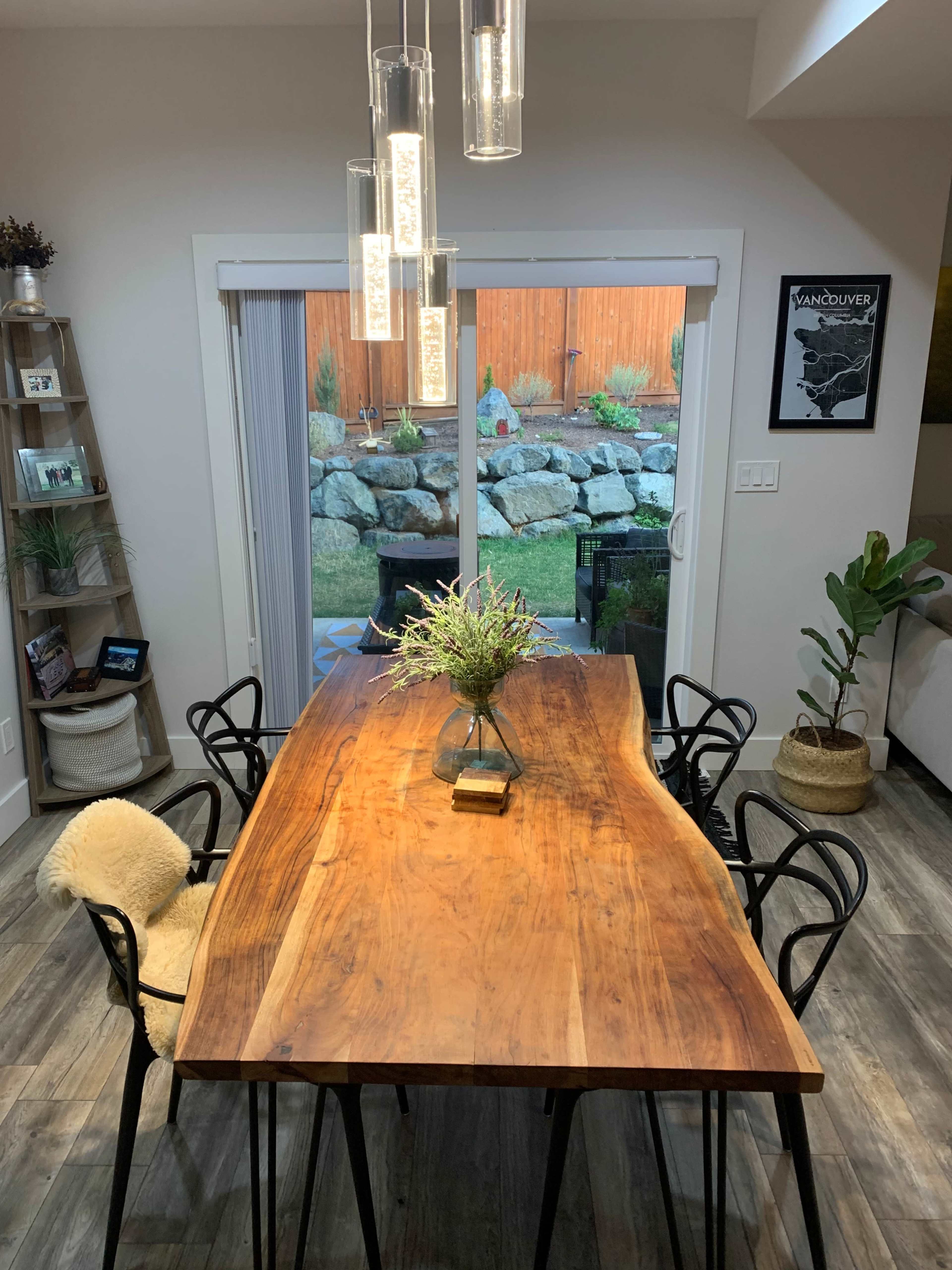 A wooden dining table with six chairs is set in a well-lit room, next to a sliding glass door that opens to a landscaped garden.