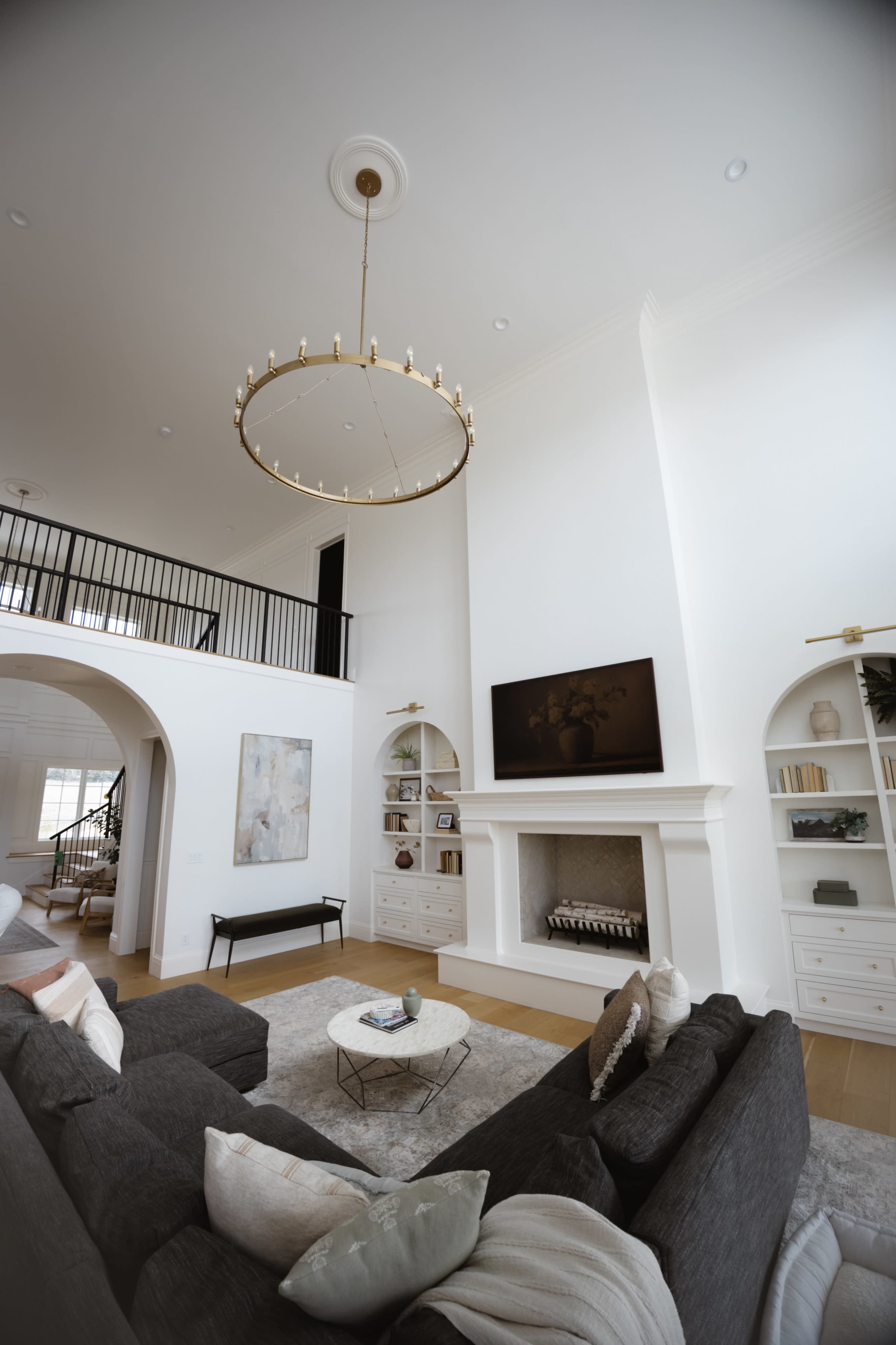 The image shows a spacious living room featuring a large circular chandelier, a modern fireplace with a TV above it, and a sectional sofa arranged on a light-colored rug.