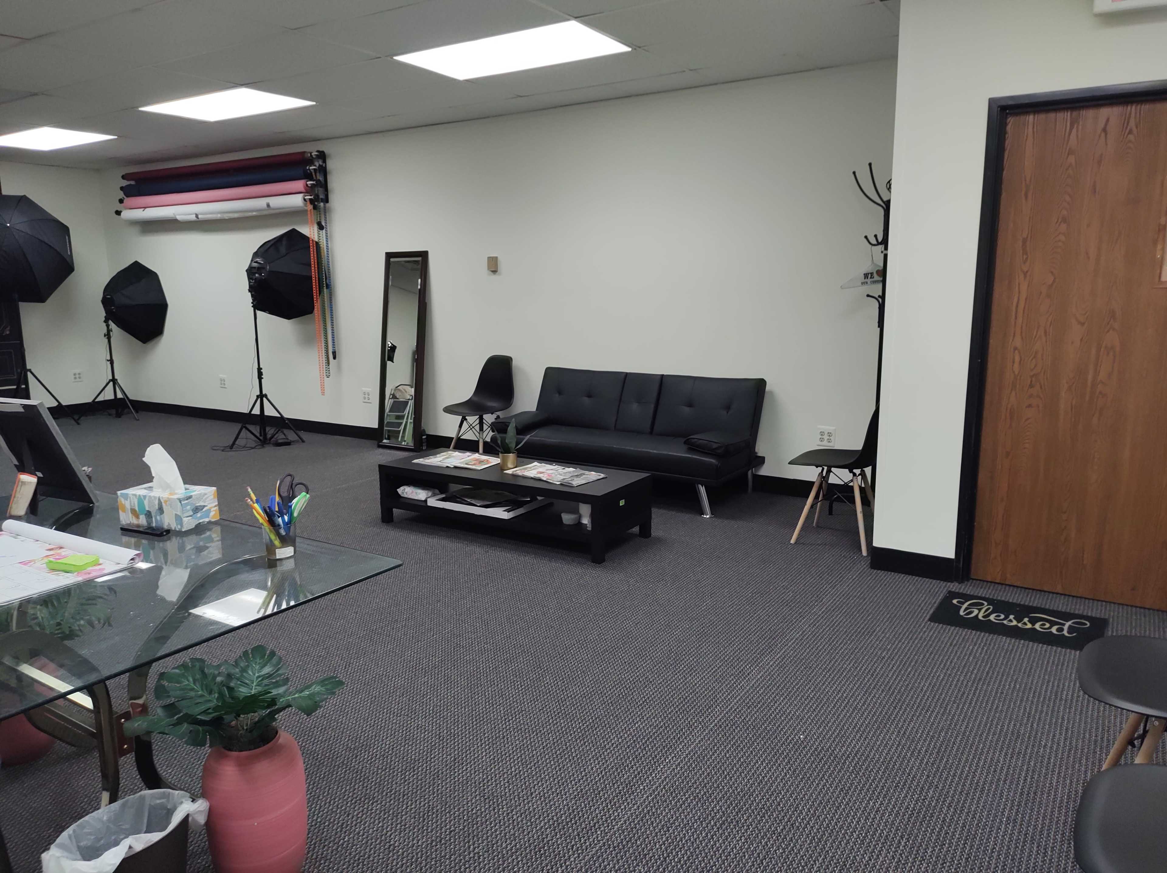 A spacious room with a black sofa, a coffee table stacked with magazines, and photography equipment, including lights and a mirror, along with a welcoming entrance mat.