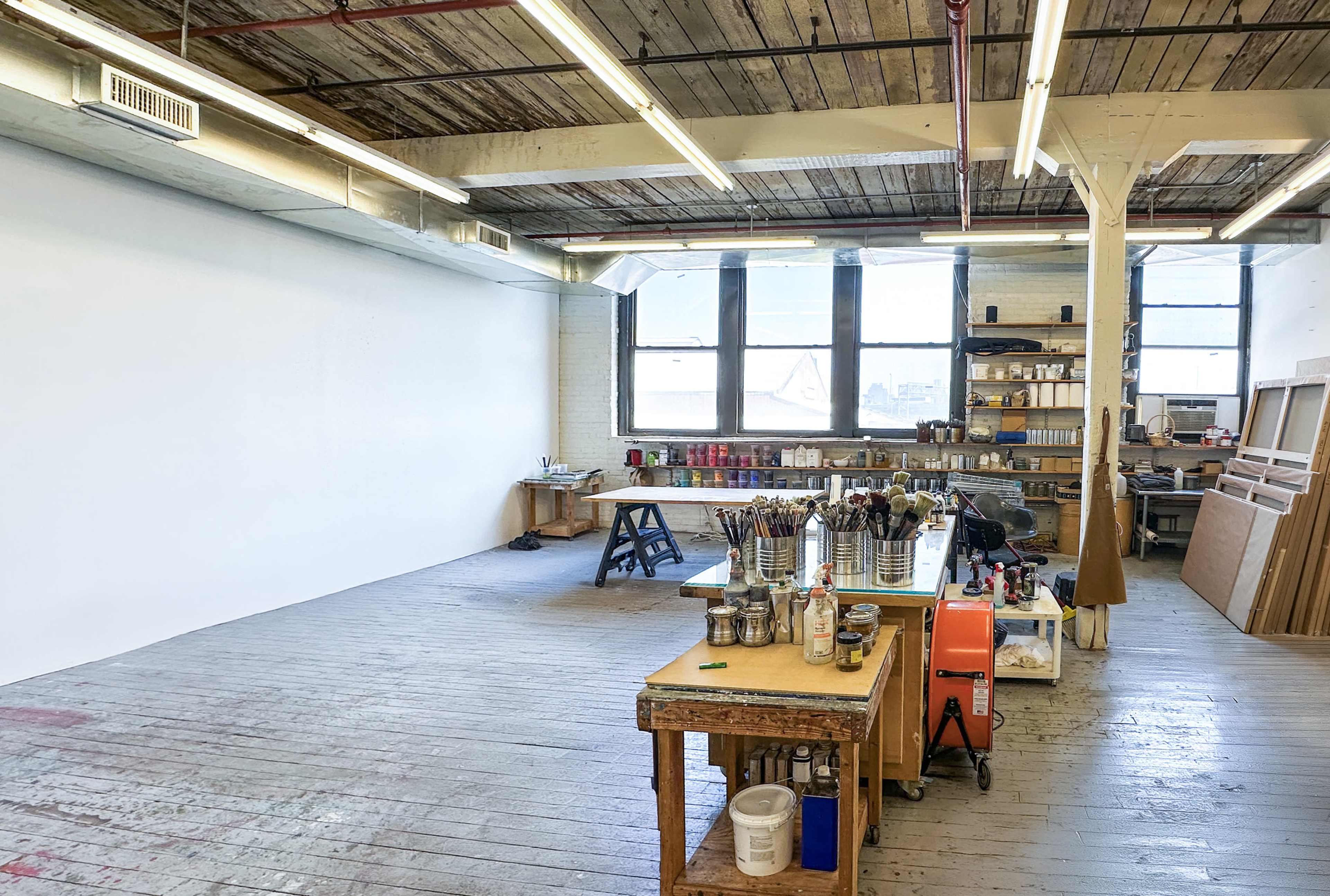 The image shows a spacious art studio with wooden floors, large windows, and various art supplies organized on tables.