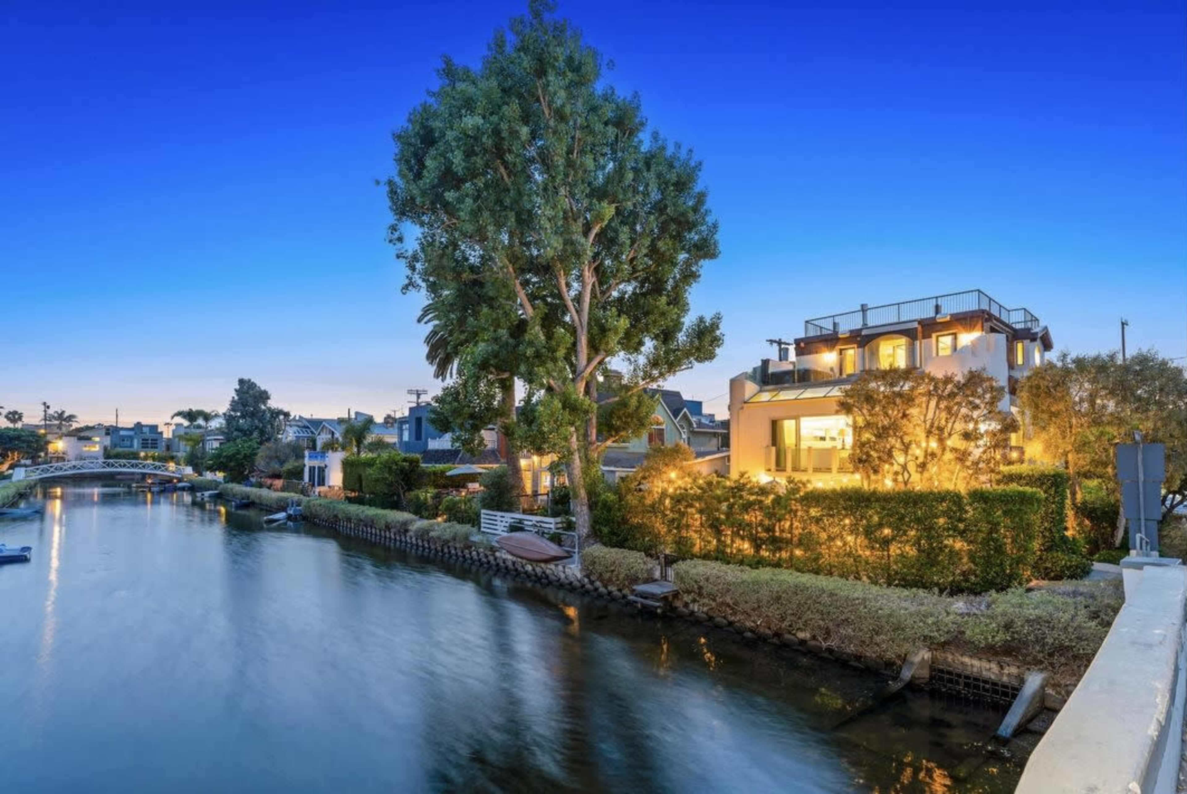 A serene canal lined with houses, illuminated in the evening twilight, with a large tree and boats visible along the water's edge.