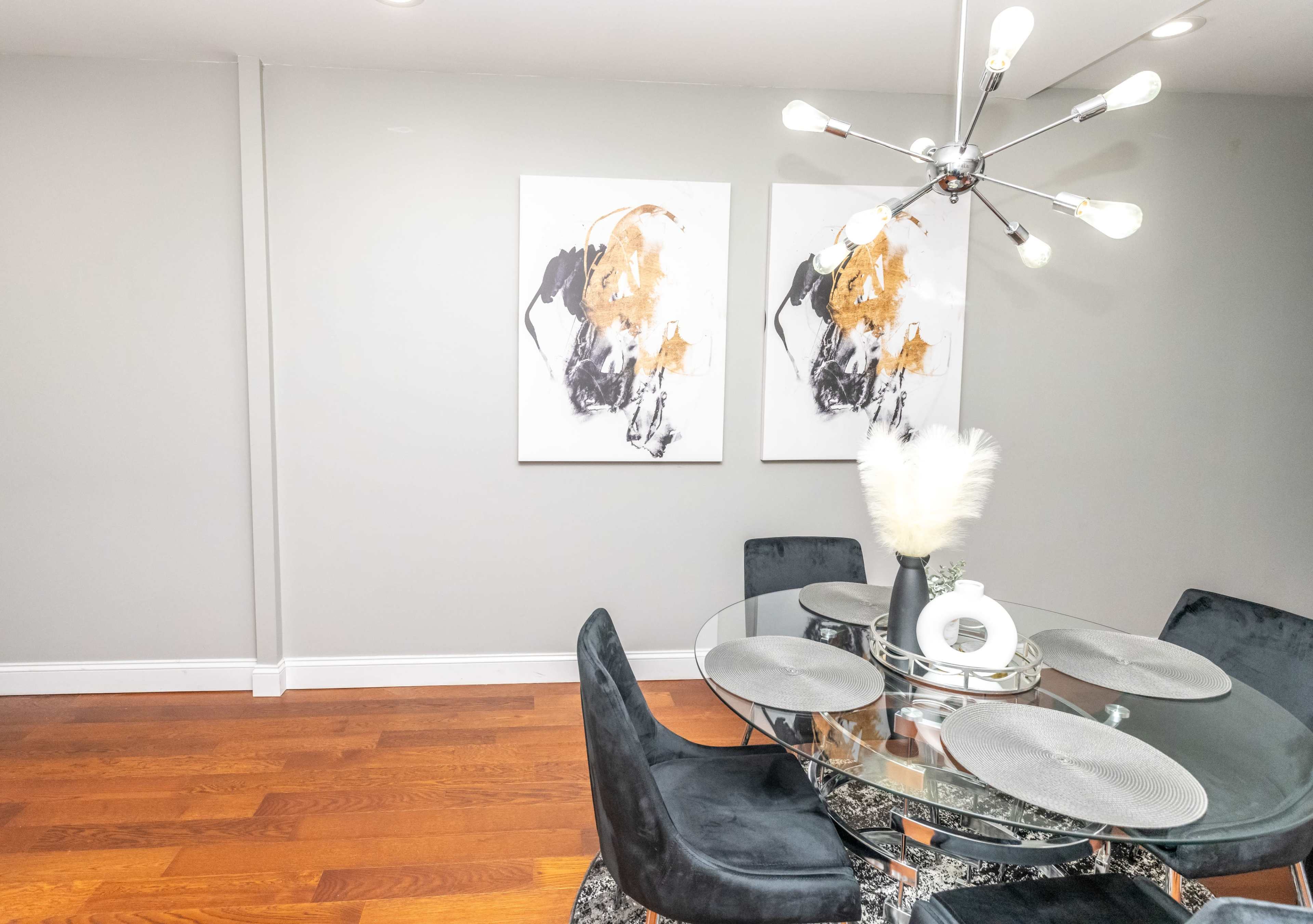 A modern dining area features a circular glass table surrounded by black upholstered chairs, with two abstract paintings on the wall and a decorative centerpiece.