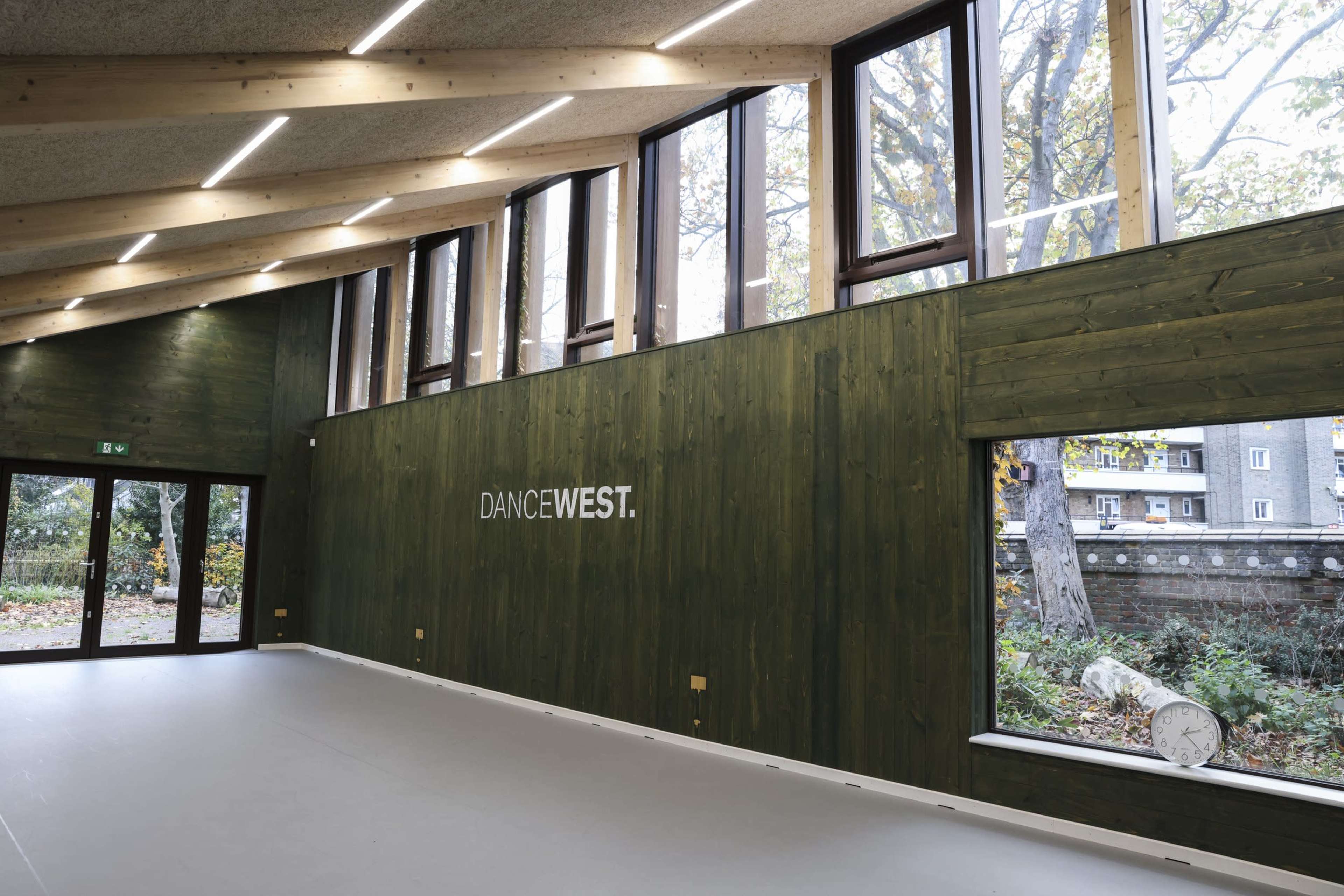 The image shows an interior space of a dance studio with large windows, wooden walls, and a minimalist design.