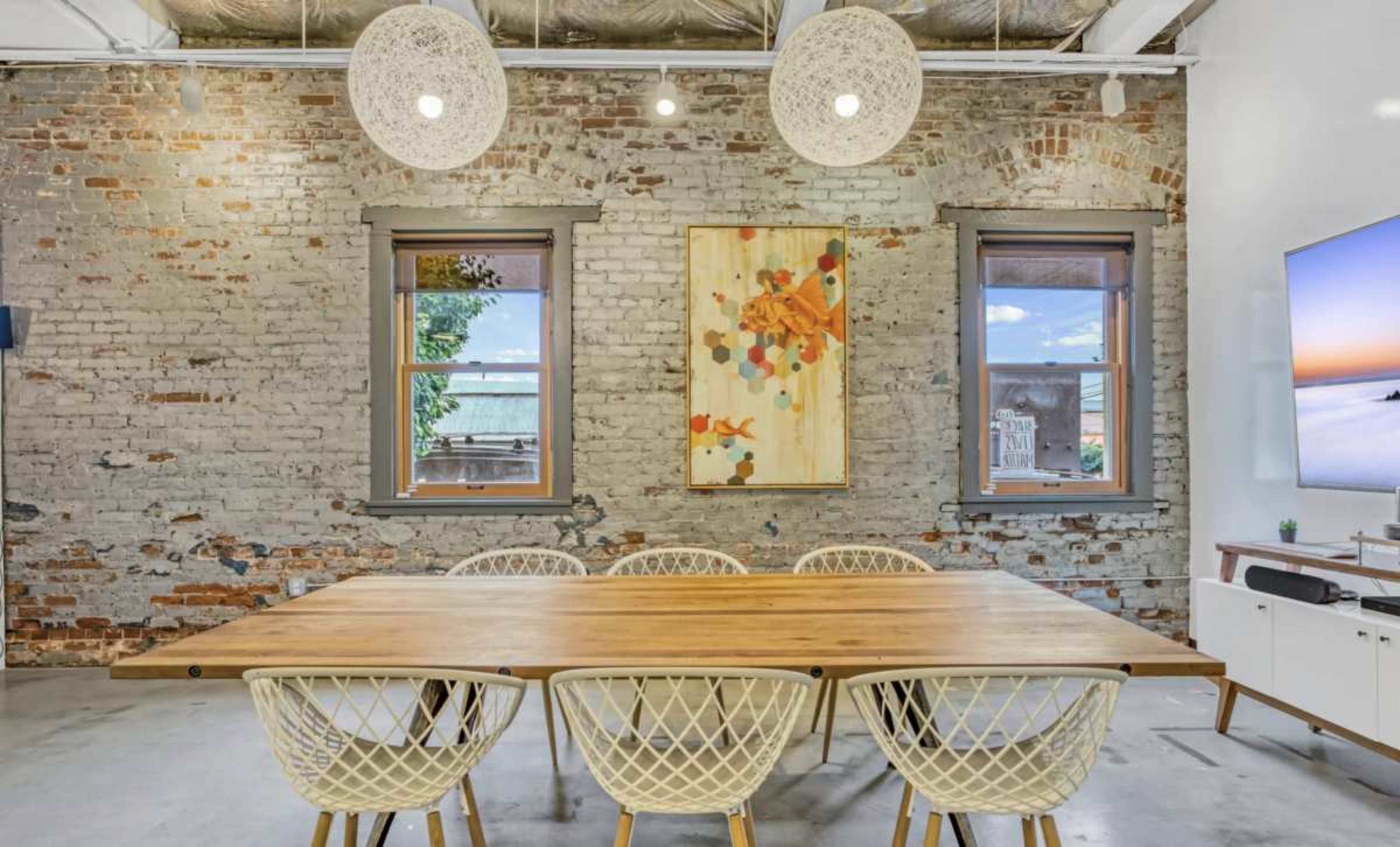 The image shows a dining area with a wooden table surrounded by six white chairs, set against a backdrop of a brick wall featuring two windows and a colorful artwork.