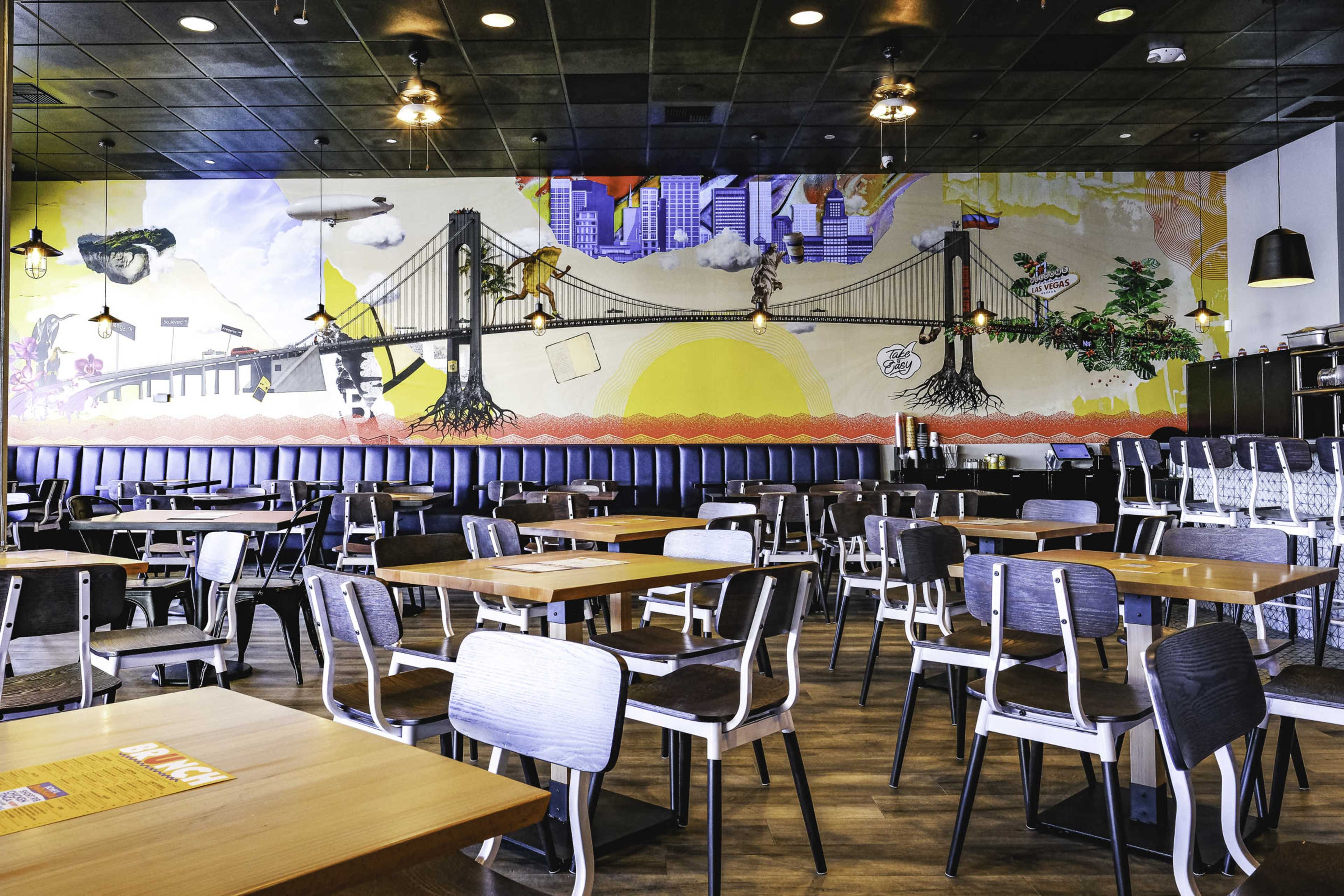 The image shows an interior of a restaurant with wooden tables and chairs, featuring a colorful mural of a bridge and cityscape on the wall.