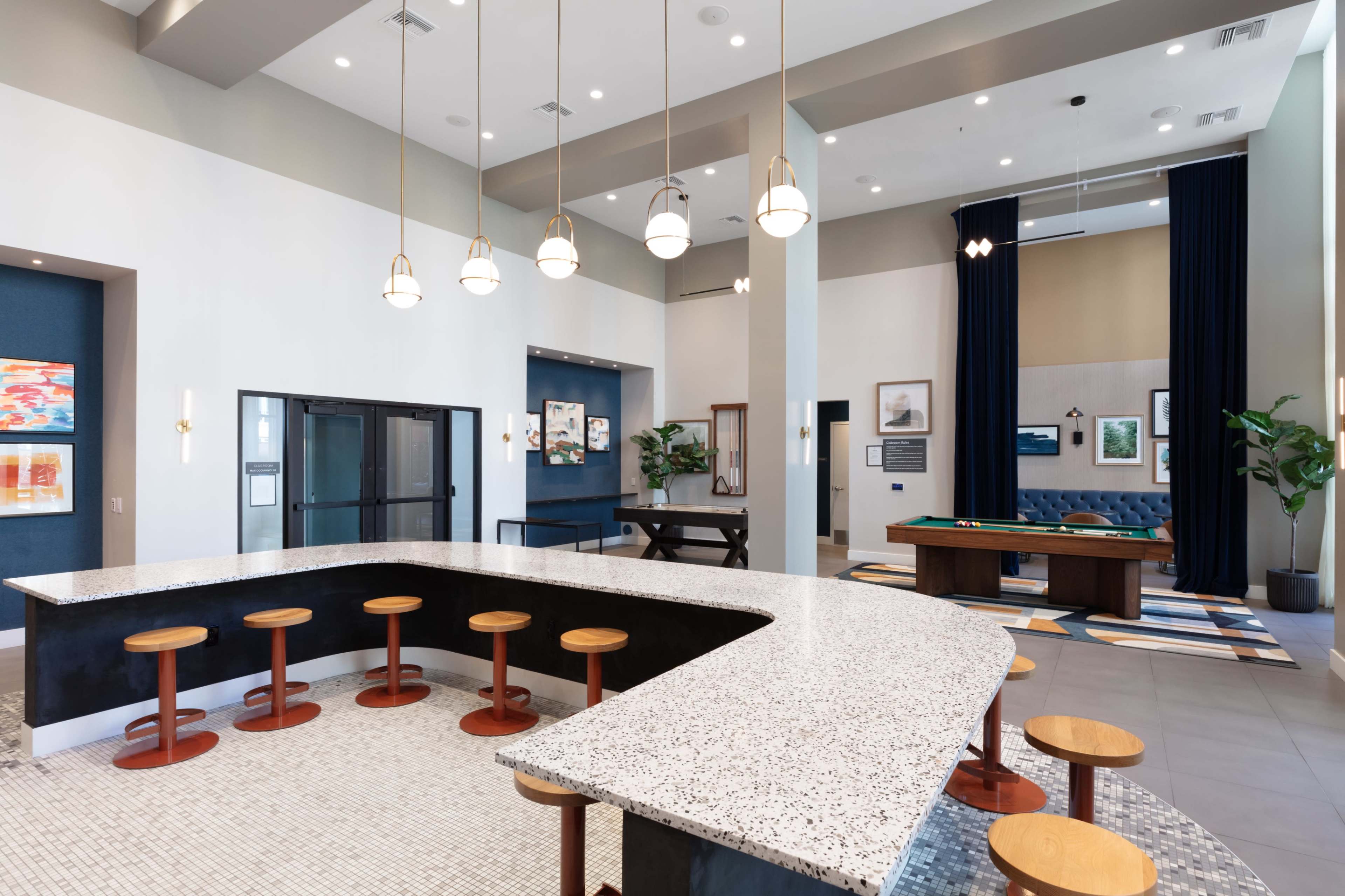 The image shows a modern communal lounge area with a large speckled countertop, wooden stools, and a billiards table, surrounded by stylish decor and ample natural light.