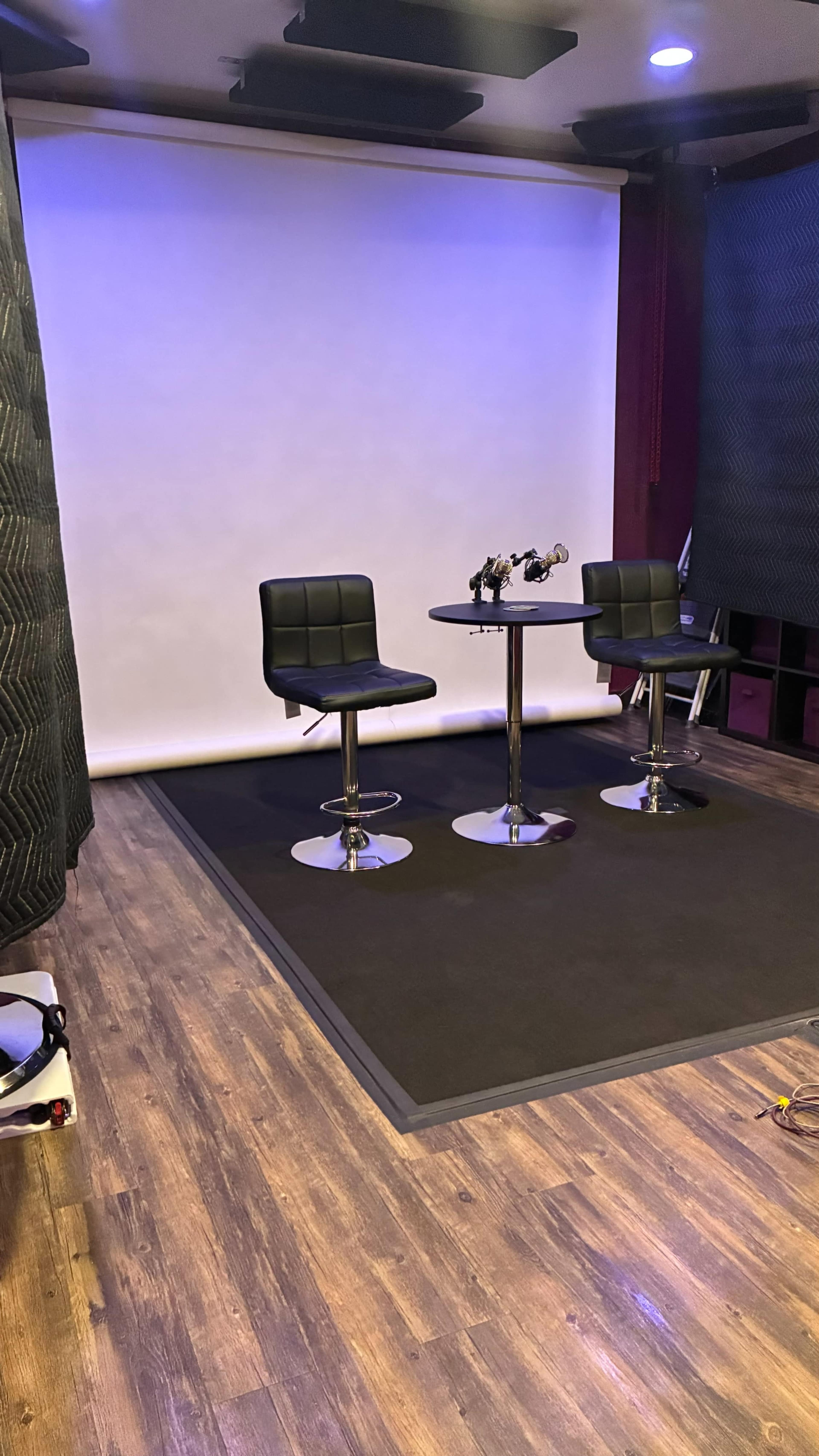 The image shows a photography or video studio featuring two black bar stools, a round table, and a blank backdrop in a well-lit room with wooden flooring.