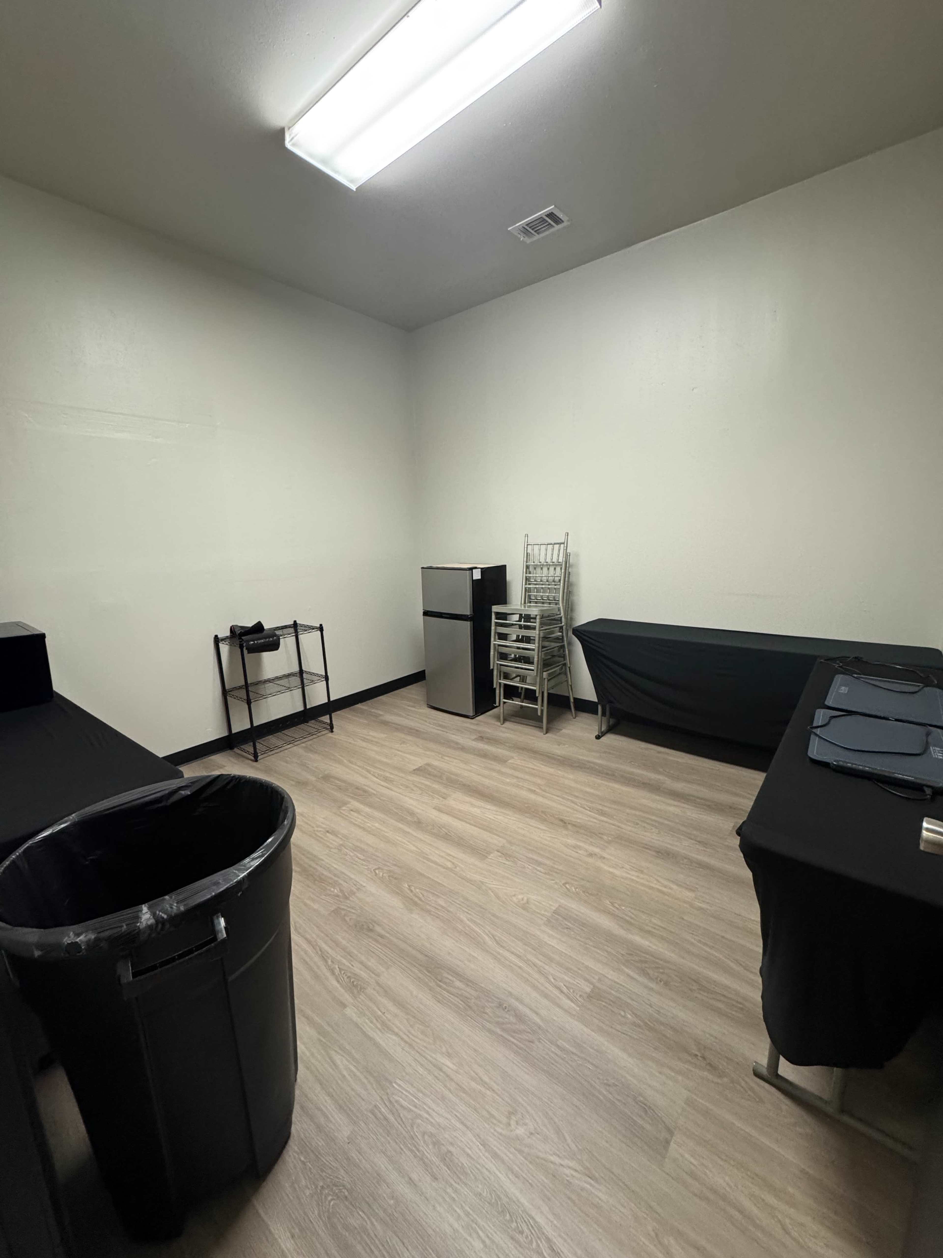 The image shows a sparsely furnished room with light wood flooring, featuring a small refrigerator, two tables covered with black cloth, a black trash can, and a stack of chairs.