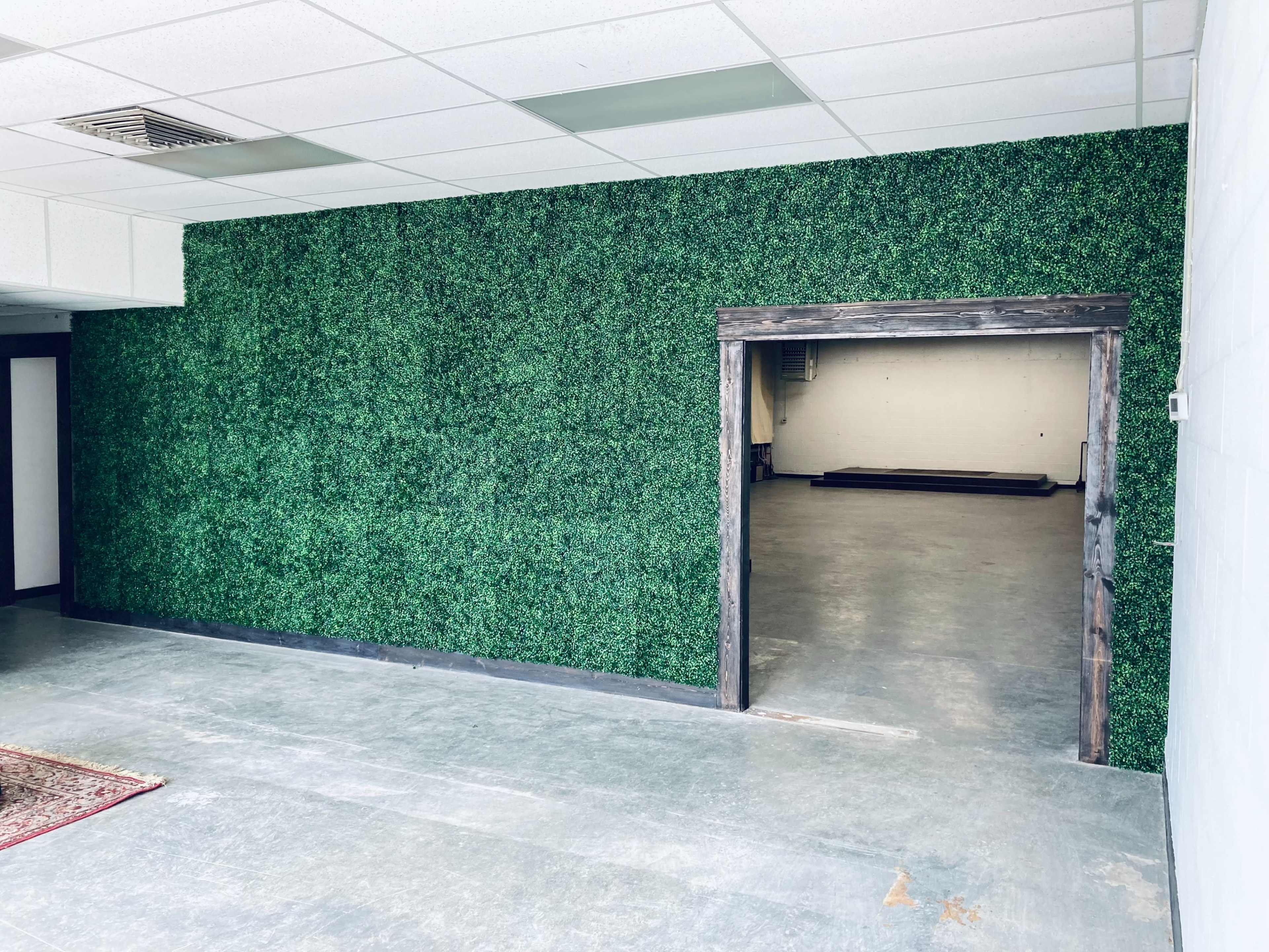 A room with a green artificial hedge wall and an open doorway leading to a space with a concrete floor and minimal furnishings.