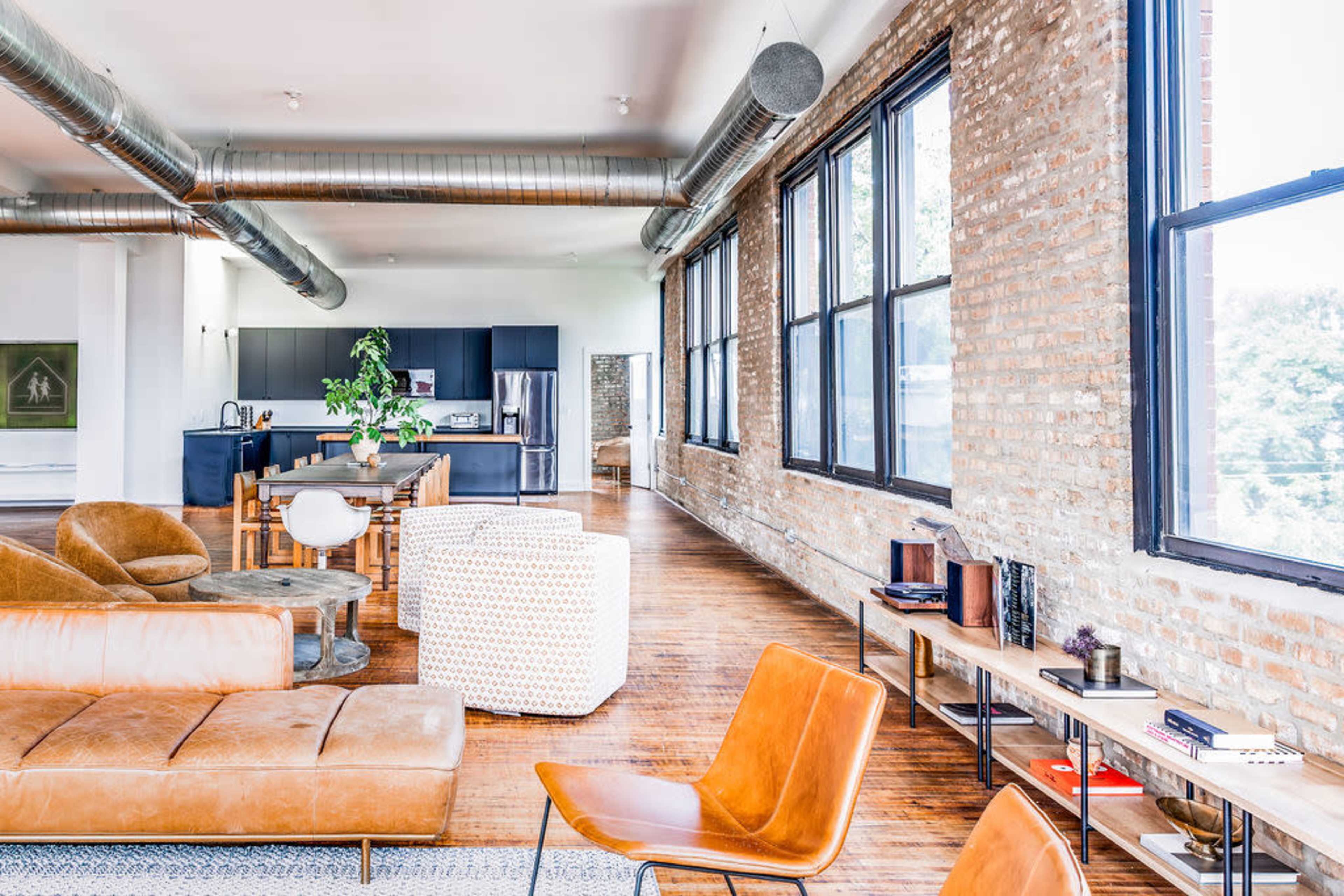 A spacious, modern living area featuring large windows, brick walls, and various seating options including a leather sofa and chairs.