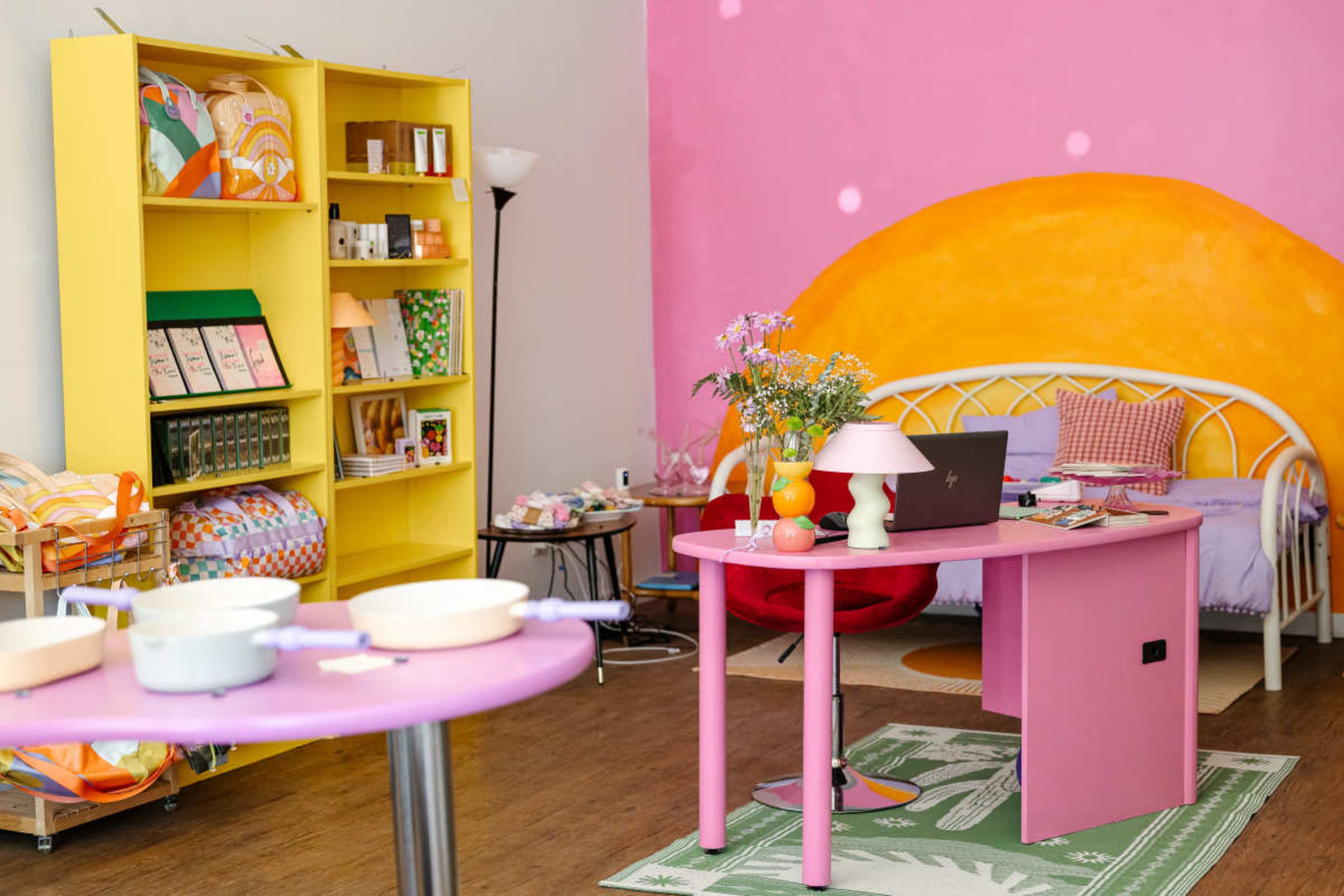 A bright, colorful room features a pink wall with an orange mural, a yellow bookshelf filled with products, a small pink table, and a cozy seating area.