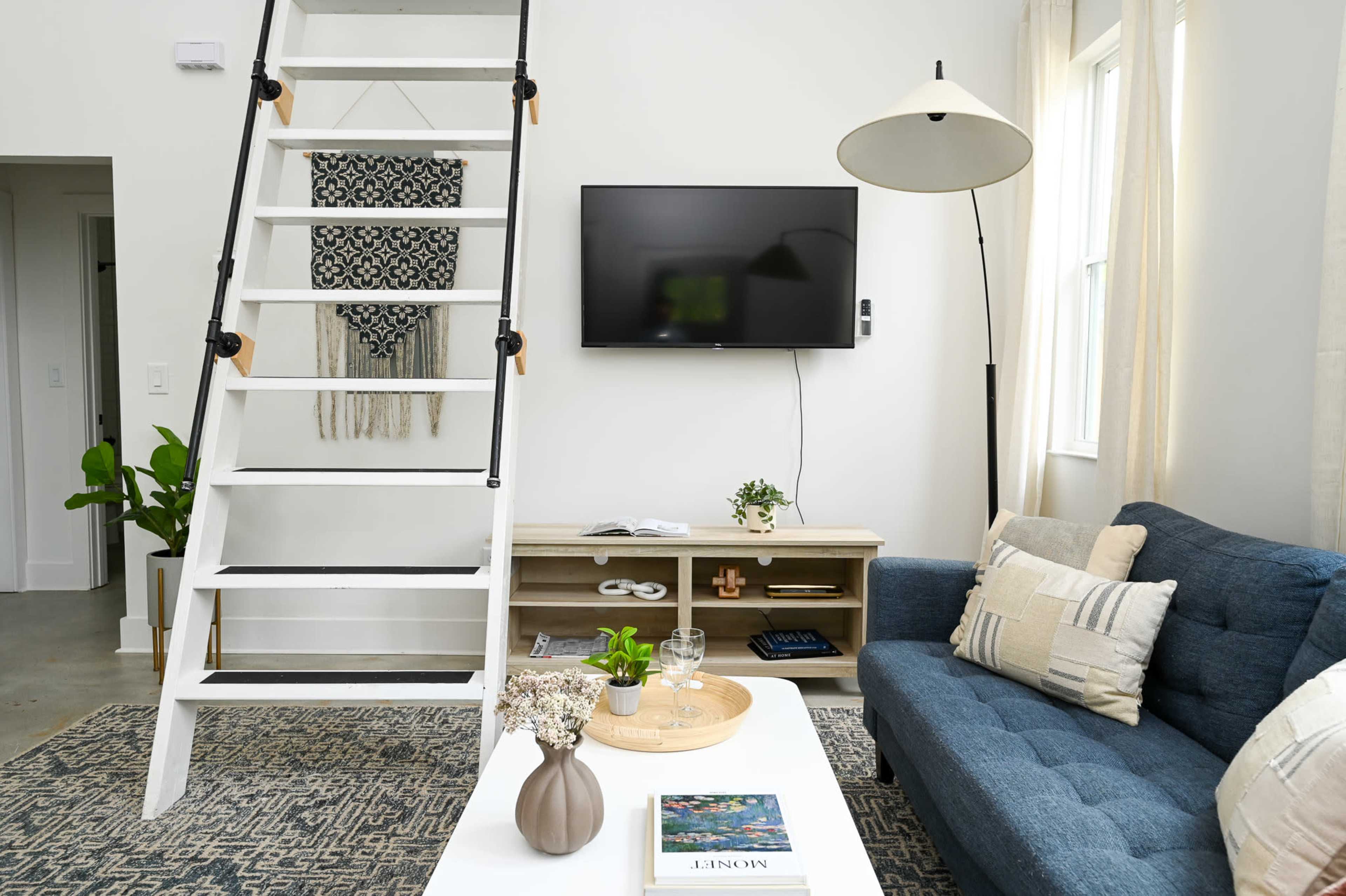 The image shows a modern living room featuring a blue sofa, a white coffee table, a television mounted on the wall, and a staircase leading to an upper level.