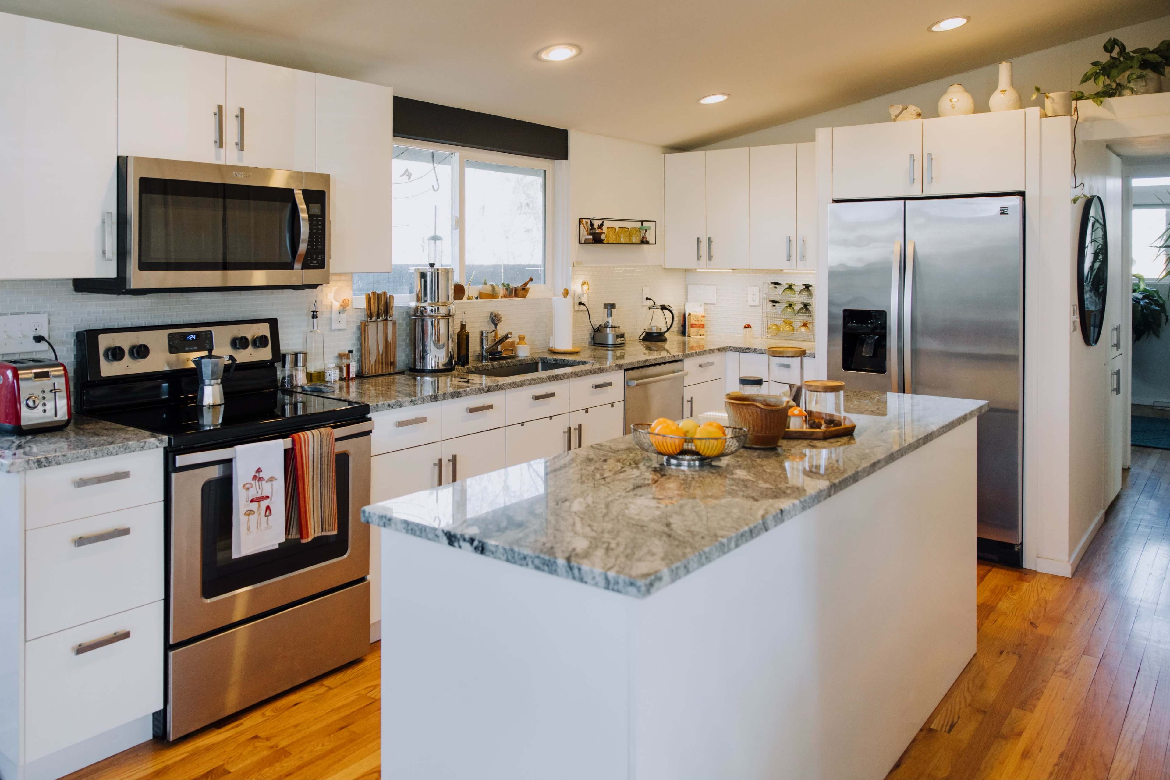 A modern kitchen features stainless steel appliances, white cabinetry, a large island with a granite countertop, and a window that lets in natural light.