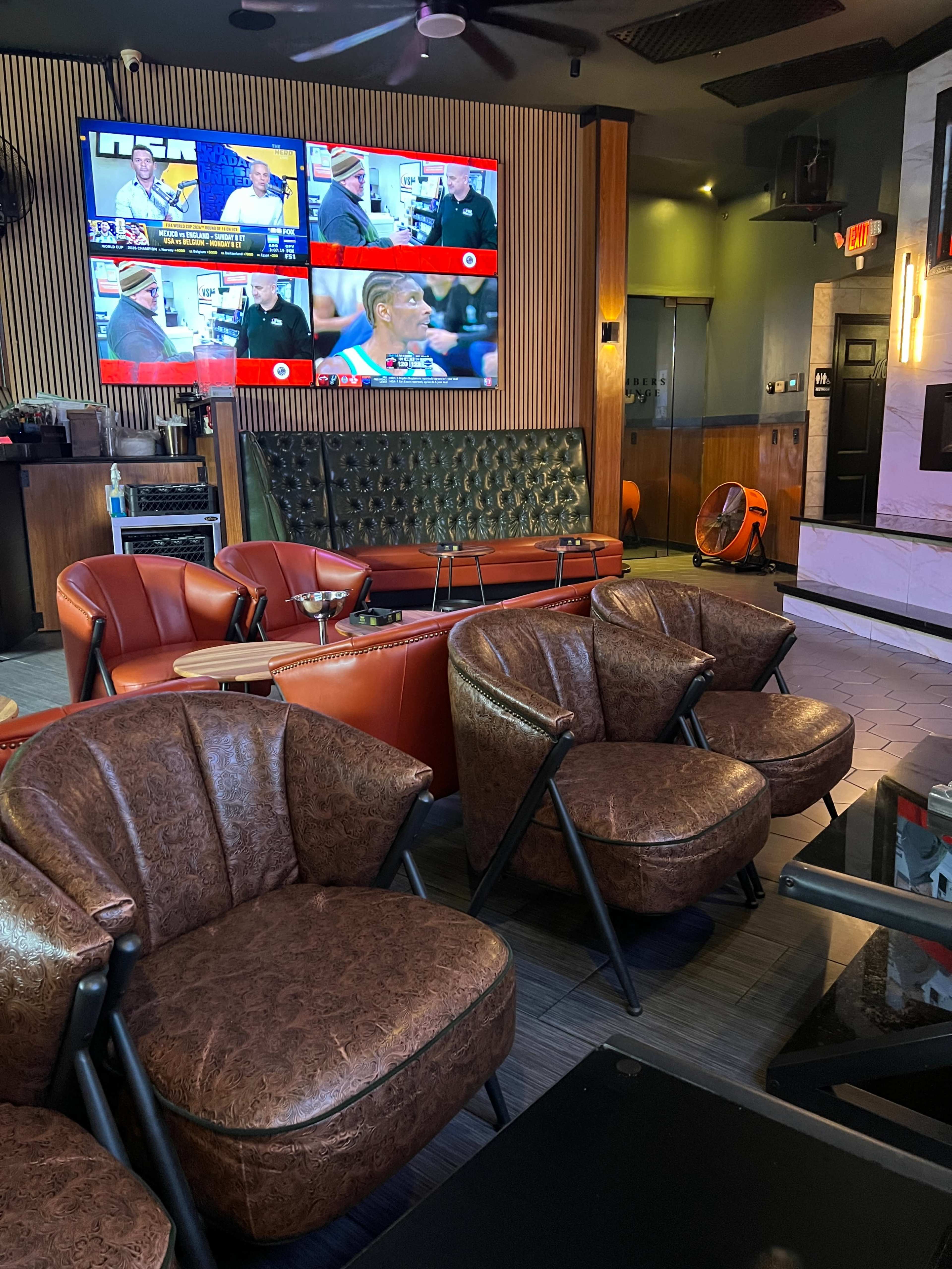 The image shows a modern lounge area with leather chairs and a large screen displaying multiple television broadcasts on the wall.