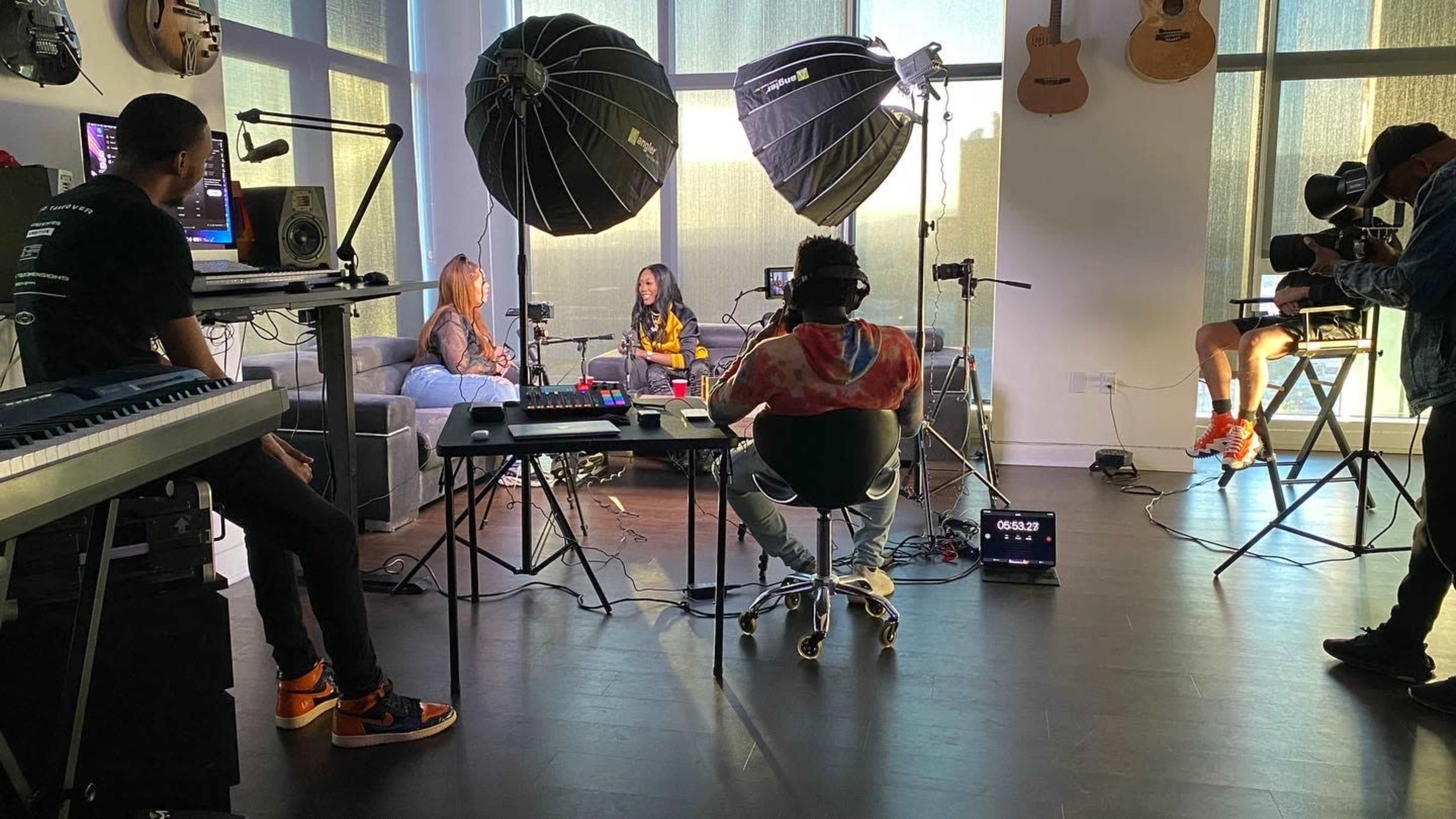 Two individuals are being filmed in a well-lit room with musical instruments on the walls and several cameras and lighting equipment set up around them.