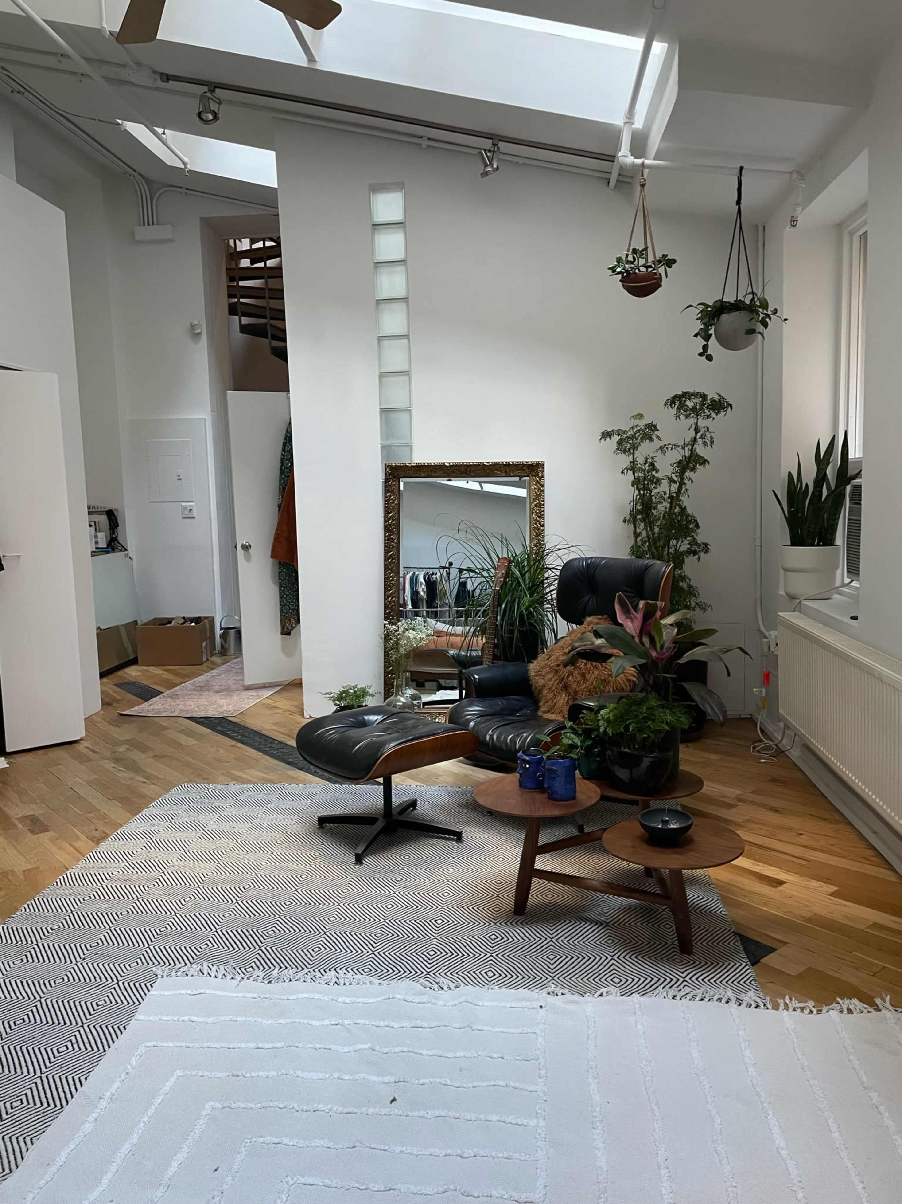 A modern living space featuring a black leather chair, a large mirror, potted plants, and a light wooden floor, all illuminated by natural light from a skylight.