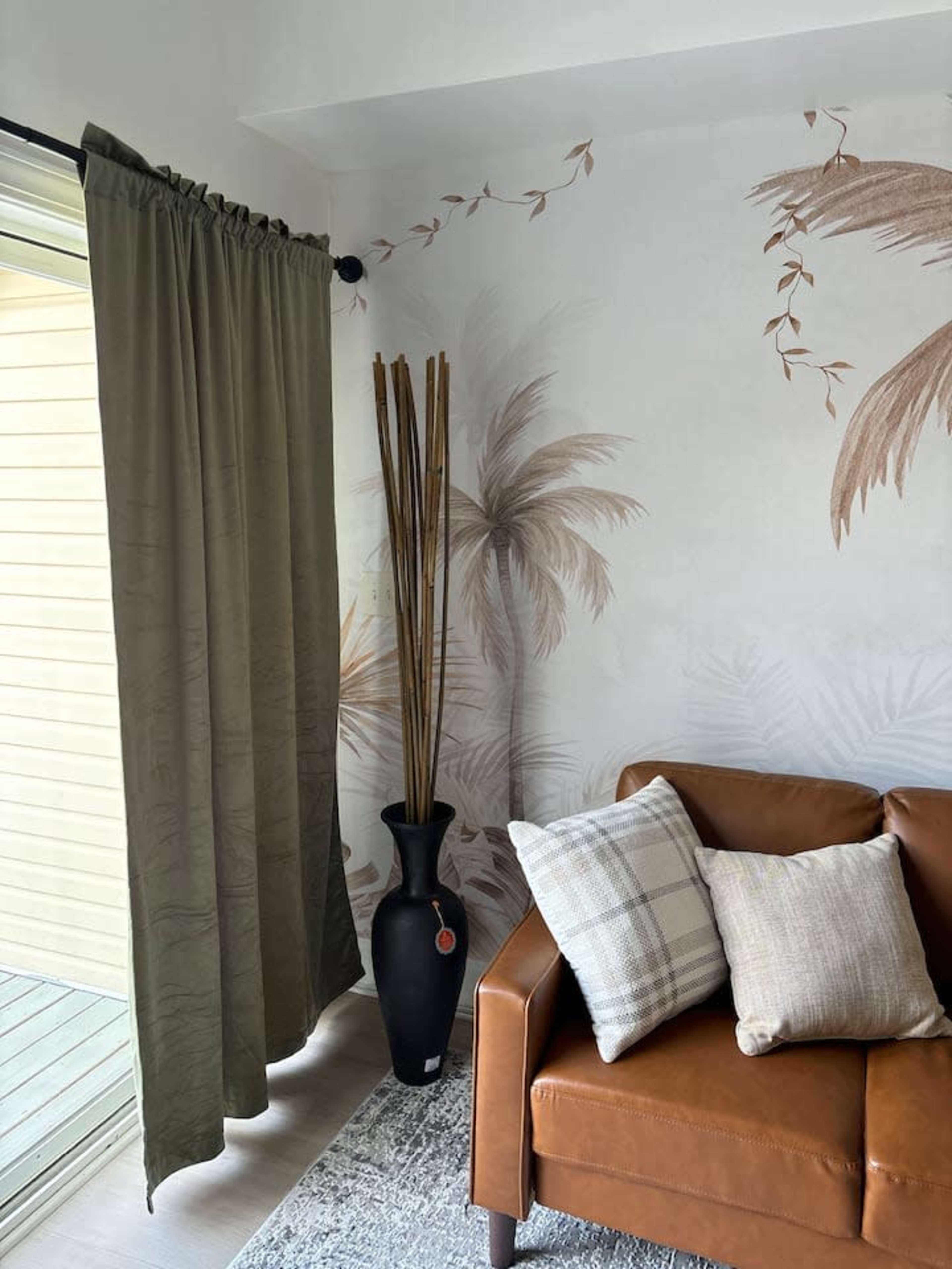 The image shows a corner of a living room with a brown leather sofa, a decorative vase with tall dried plants, and green curtains against a wallpaper featuring palm leaves.