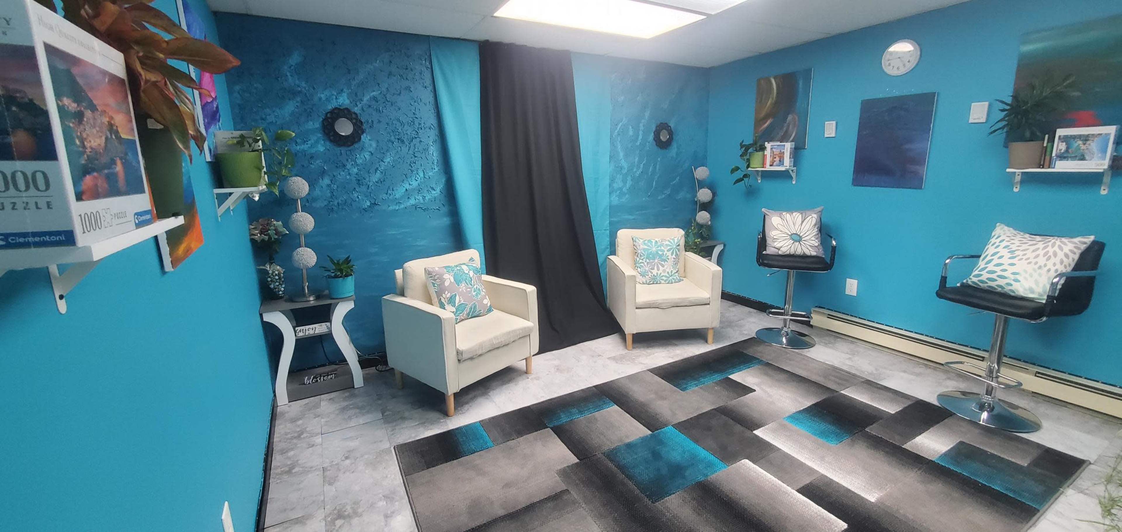 A blue-walled room with two armchairs and a black curtain, featuring decor items such as cushions, plants, and a rug with geometric patterns.